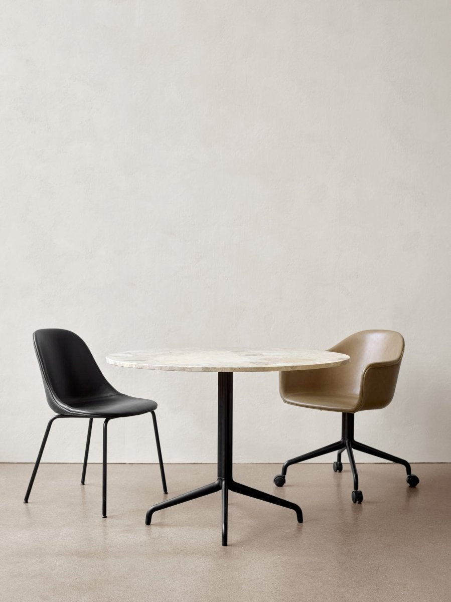 Round marble-top dining table with black metal pedestal base flanked by a black leather side chair and tan leather swivel