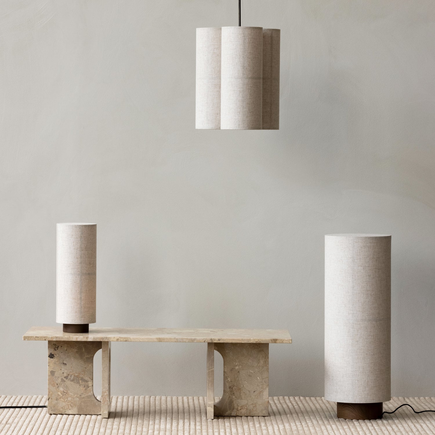 Beige cylindrical fabric pendant light and matching table lamps over travertine bench, neutral textured backdrop