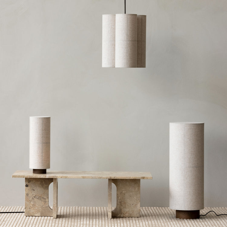 Beige cylindrical fabric pendant light and matching table lamps over travertine bench, neutral textured backdrop