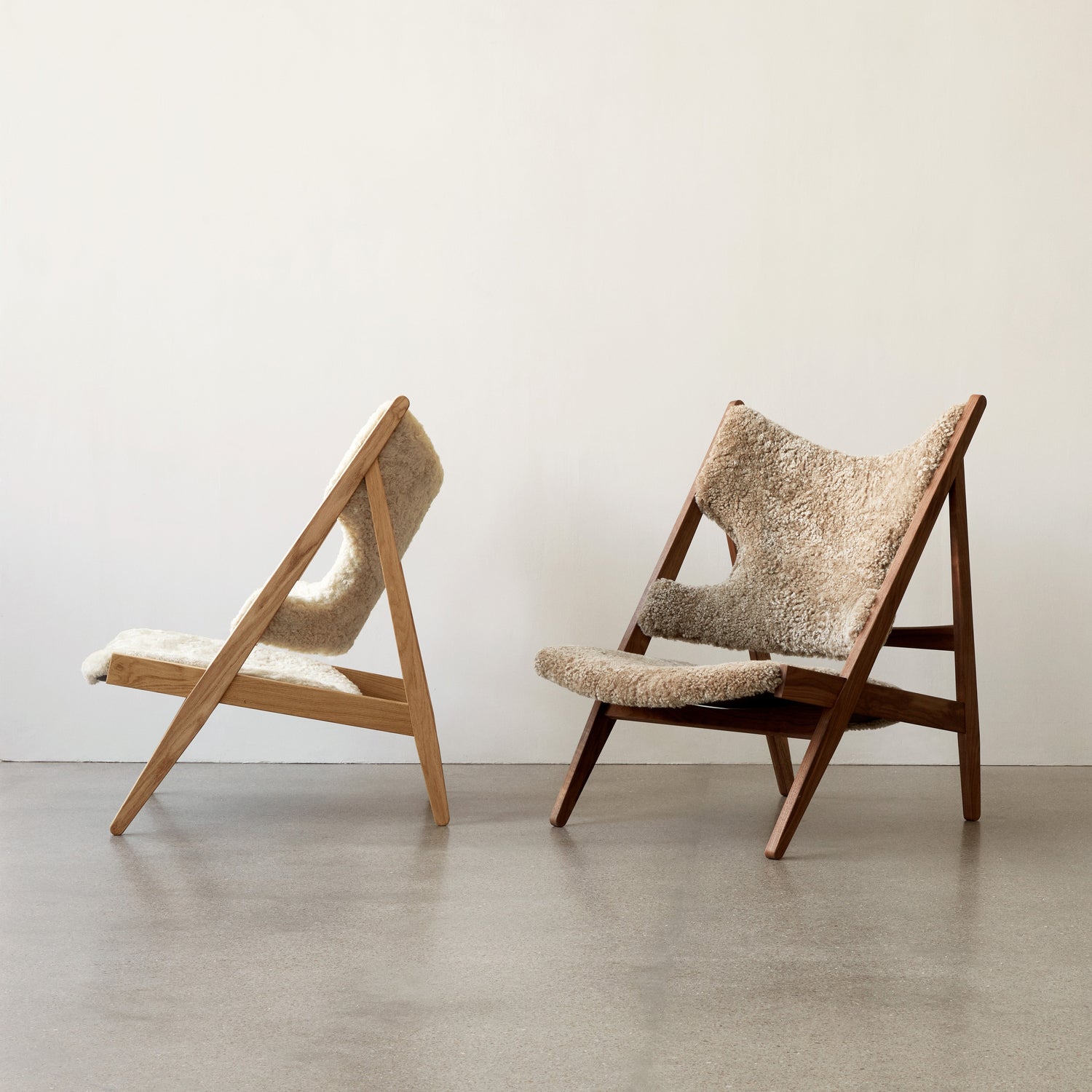 Two lounge chairs with angled oak frames and cream sheepskin upholstery against a pale wall.
