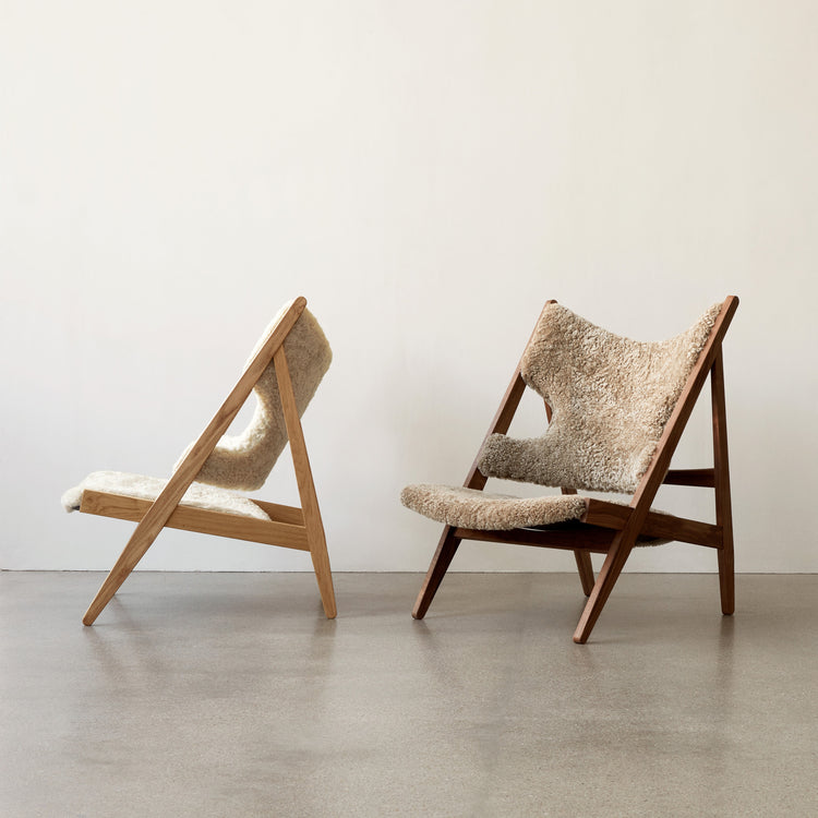 Two lounge chairs with angled oak frames and cream sheepskin upholstery against a pale wall.