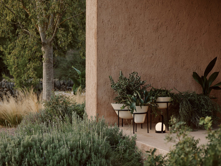 Outdoor beige ceramic plant pots on black metal stands with green foliage, placed on a terracotta textured porch with soft
