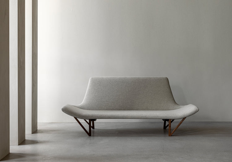 Minimal grey upholstered two-seater sofa with curved seat and wooden V-shaped legs against a pale concrete wall.