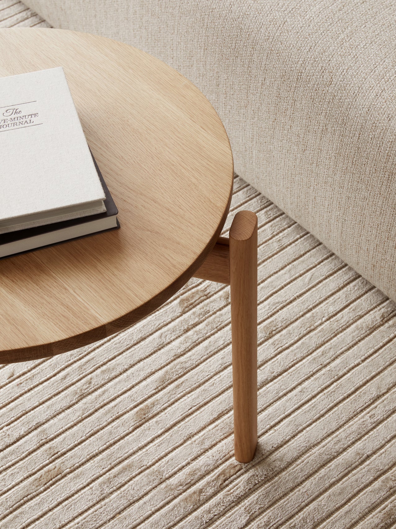 Close-up detail of the wooden Passage Lounge Table with books, seen on the striped Houkime Rug next to a beige sofa.