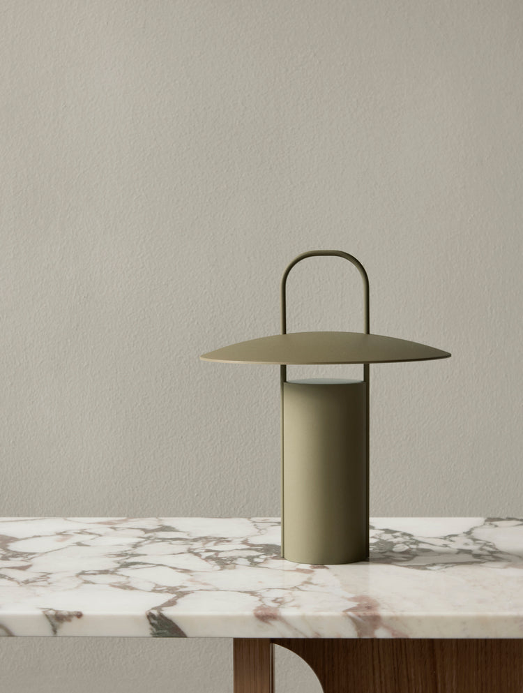 Olive-green matte metal table lamp with cylindrical base, wide disc shade and arched handle on marble tabletop