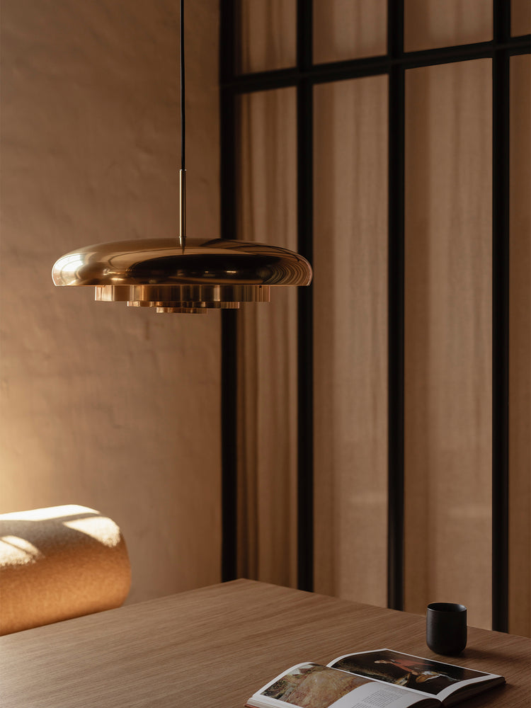 Brass dome pendant light with polished finish hanging above a wooden table with open book and black cup, warm beige tones