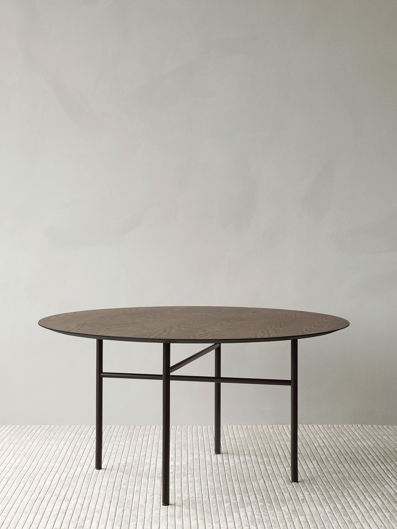 Round dark oak table top with thin black metal legs and crossbar base on a textured light rug against pale wall