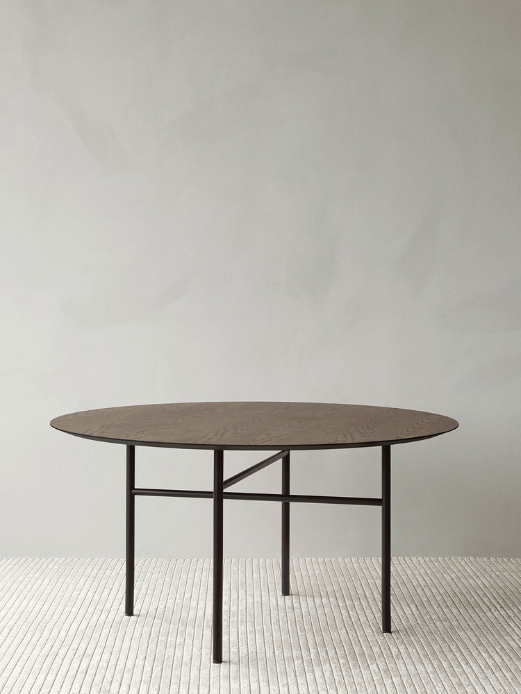 Round dark oak table top with thin black metal legs and crossbar base on a textured light rug against pale wall
