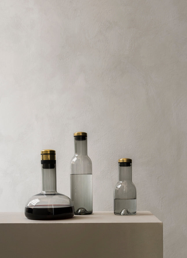 Three smoked-glass bottles with brass lids—one wide decanter and two tall slender carafes—on a pale plinth against textured