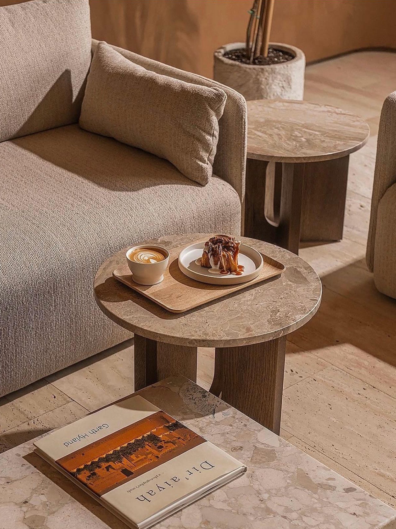 Beige textured sofa with lumbar cushion beside round stone-top side table holding latte and caramel pastry on a wooden tray