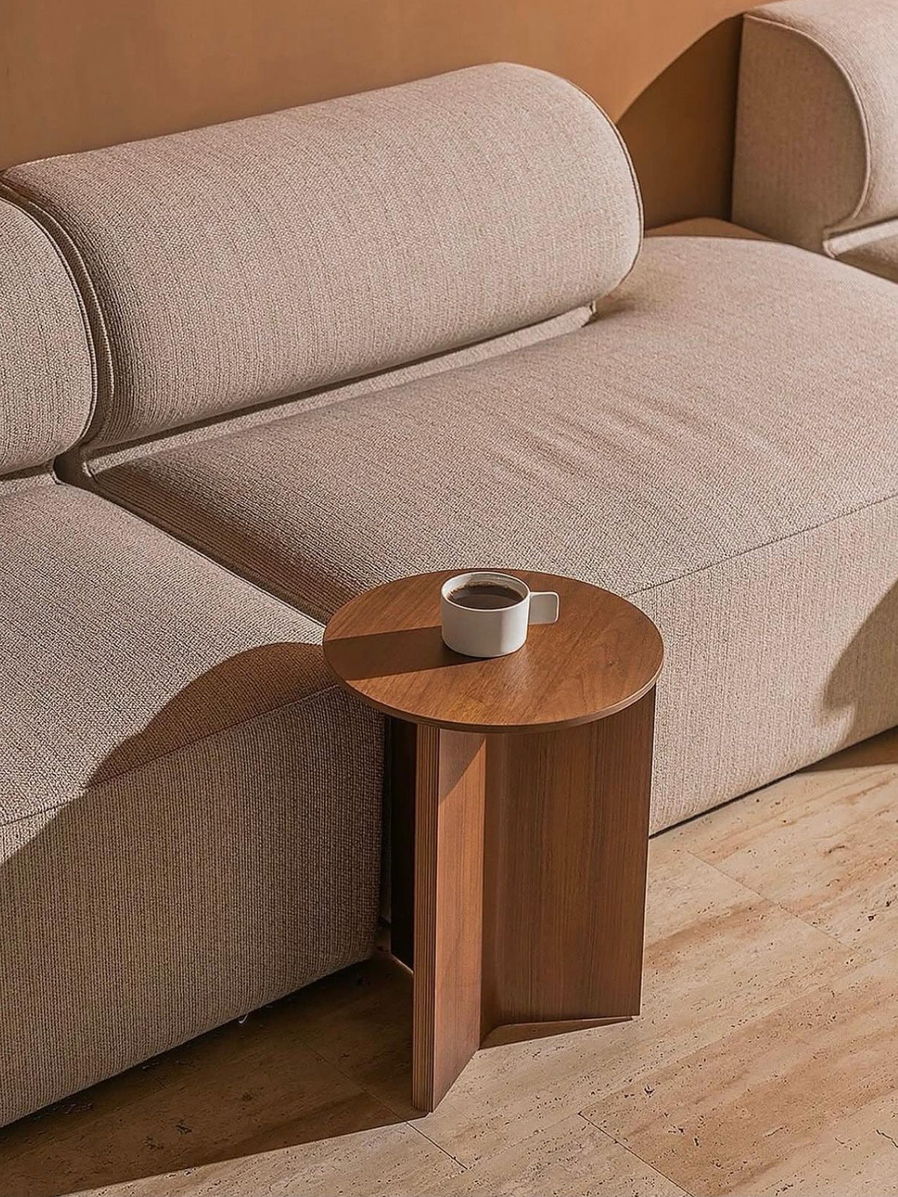 Beige textured fabric sofa with rounded bolstered backrests and a small wooden side table holding a white coffee cup