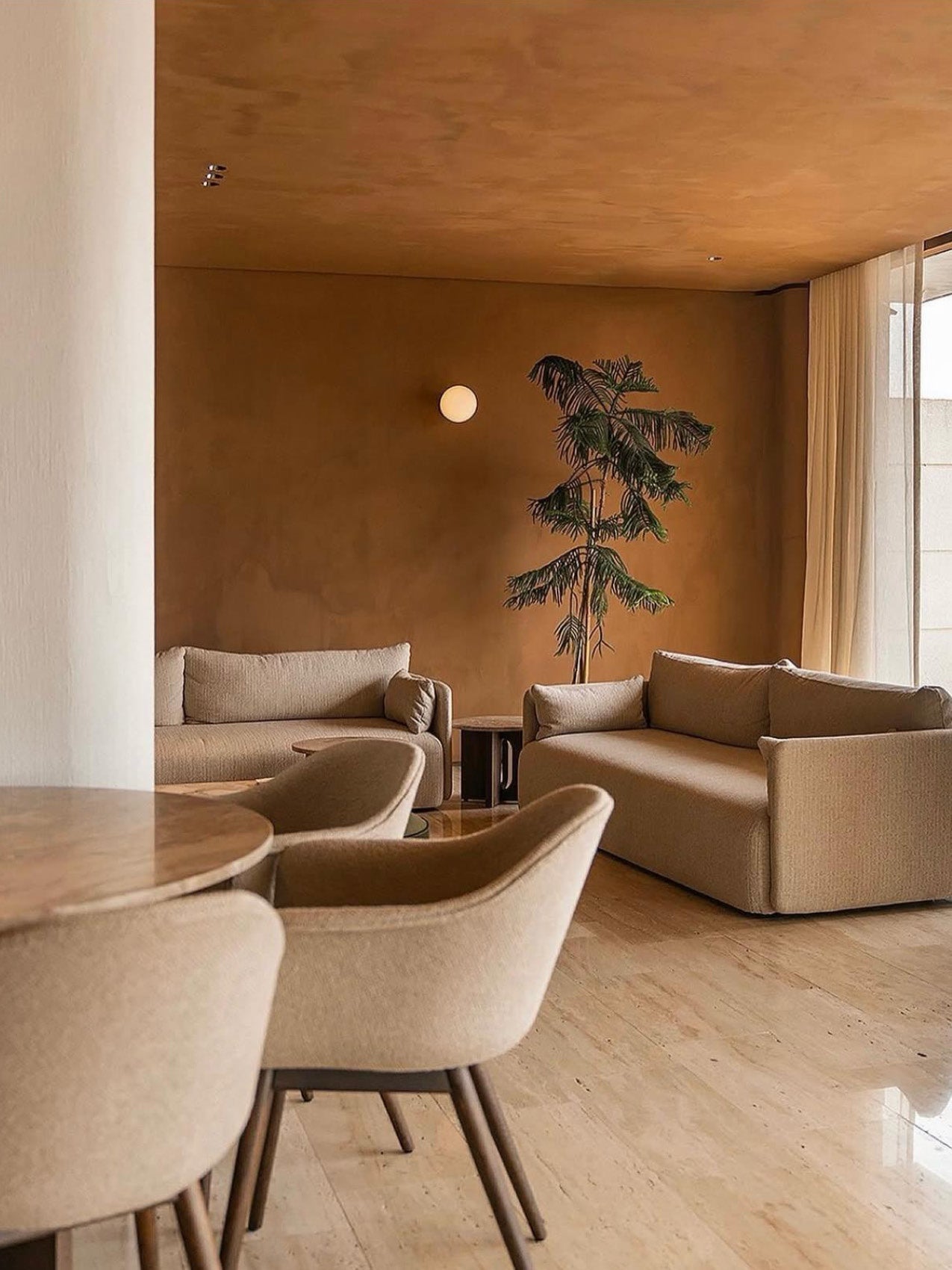 Warm-toned living room with beige upholstered sofas and chairs, marble floor, round wall light and tall potted palm.