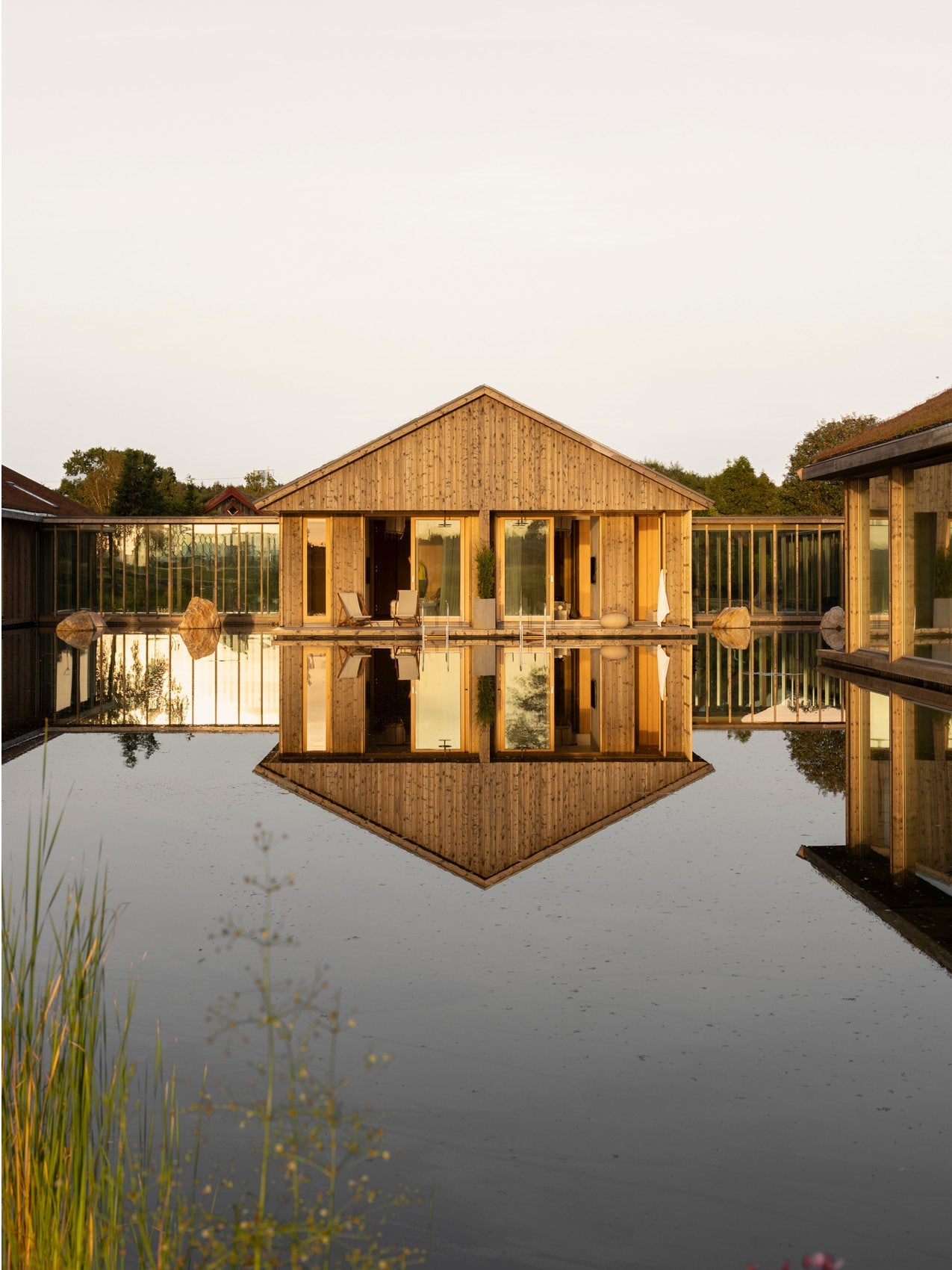 Wooden gabled pavilion with large glass doors reflected symmetrically in calm black water, warm golden timber and stone