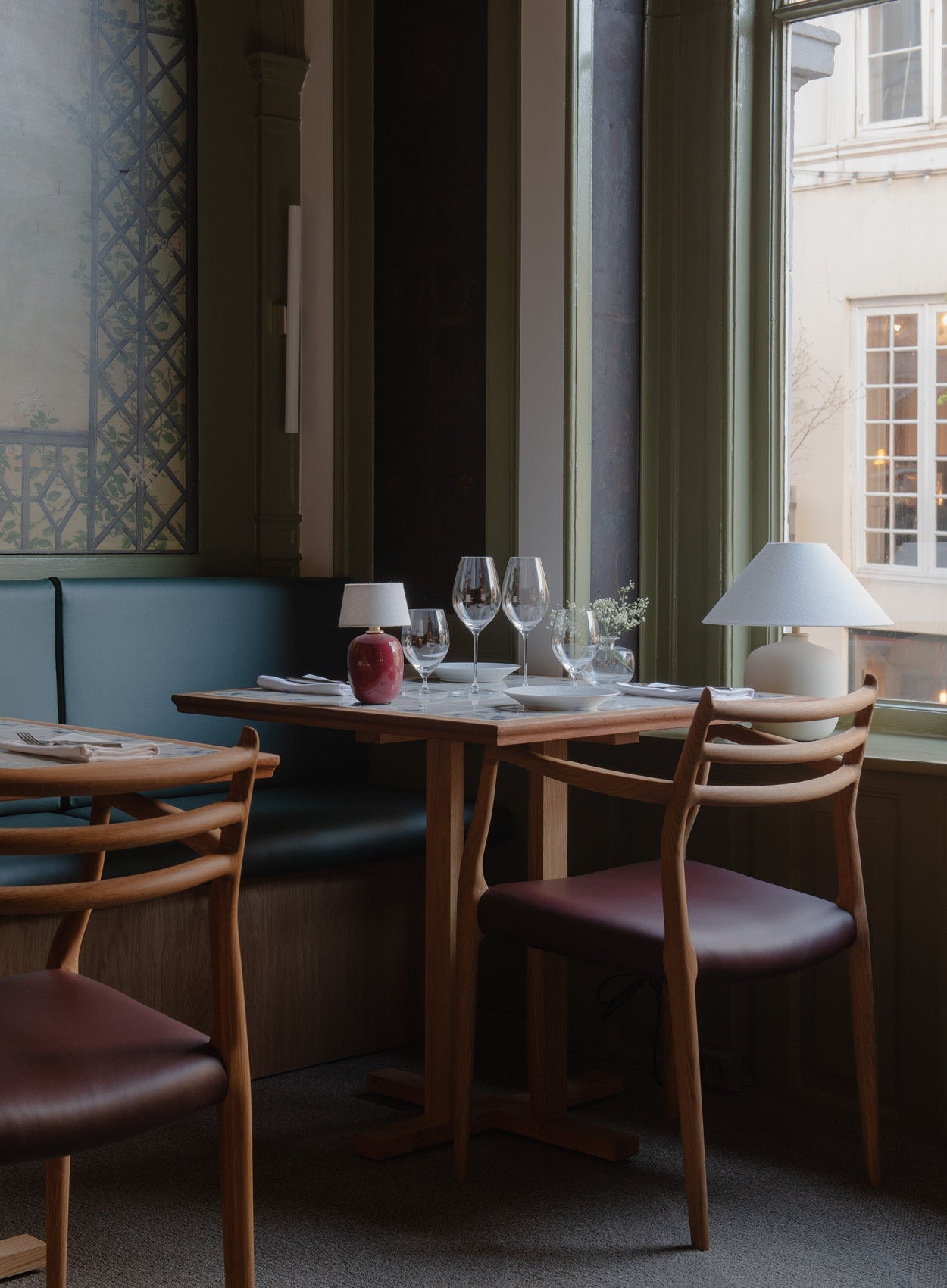Wood dining table set with wooden chairs, leather seats, two ceramic table lamps, wine glasses and place settings by a window