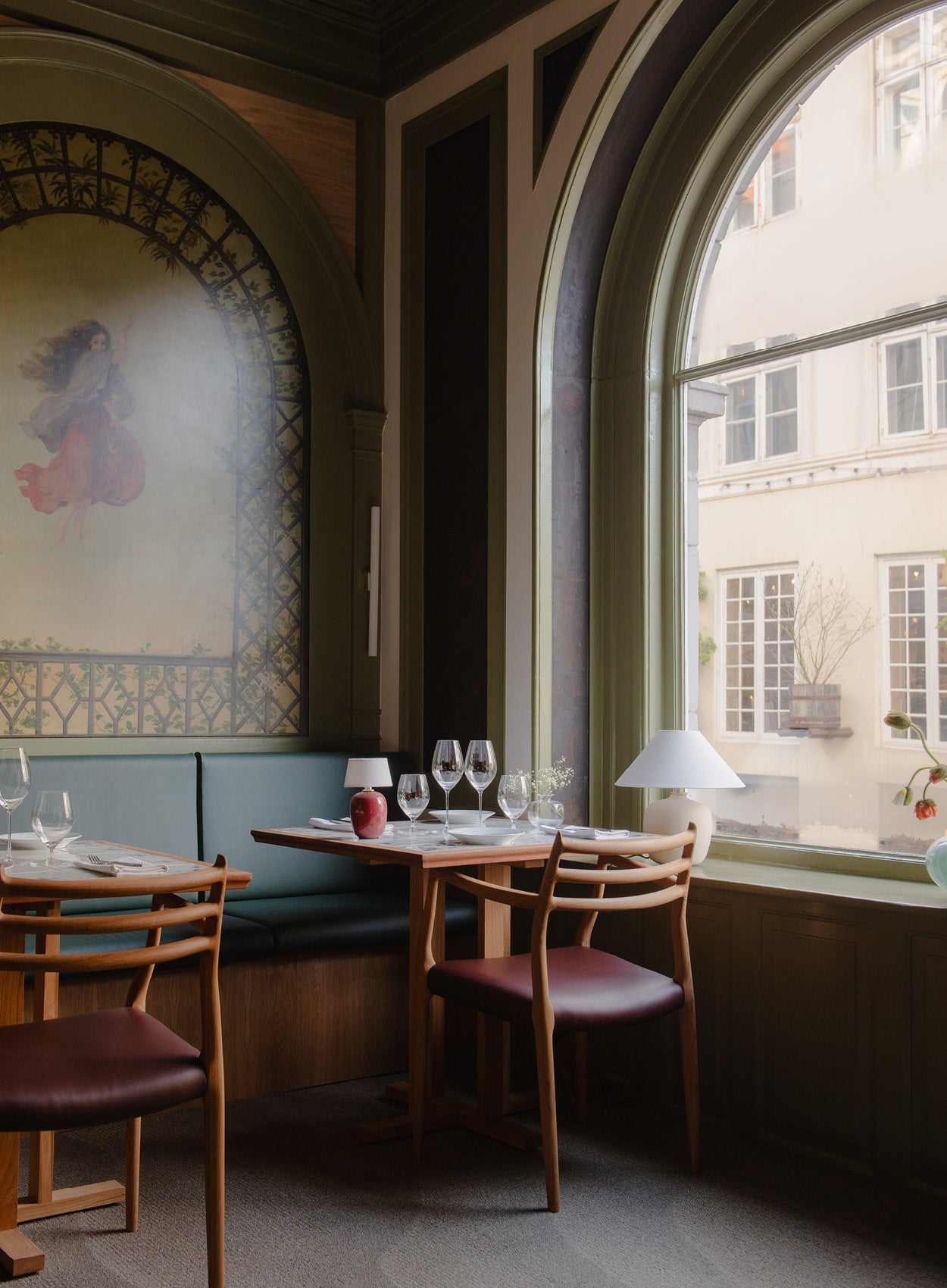 Cozy restaurant corner with wooden table, teal banquette, curved arched window, wine glasses and small white table lamp
