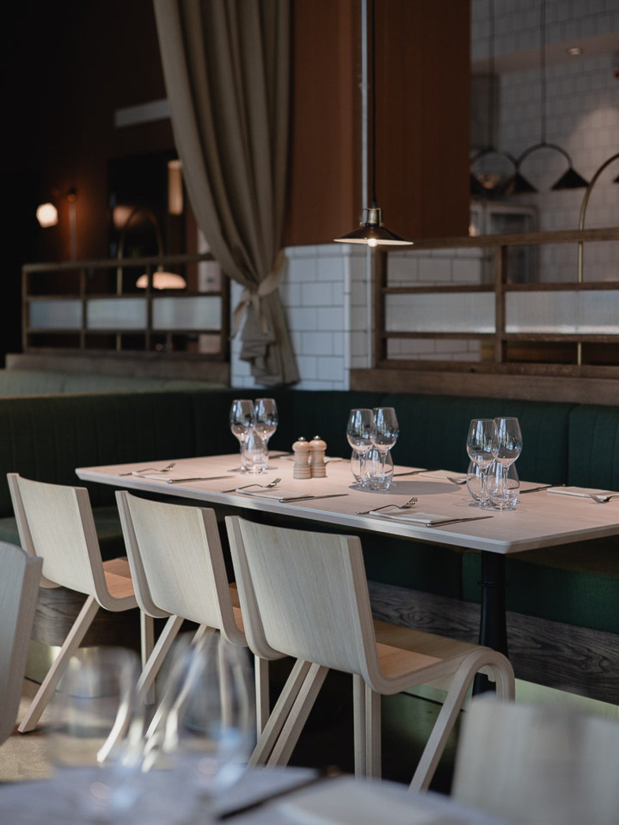 Minimal light wood dining chairs and table set with glassware, salt and pepper shakers, dark green banquette and hanging