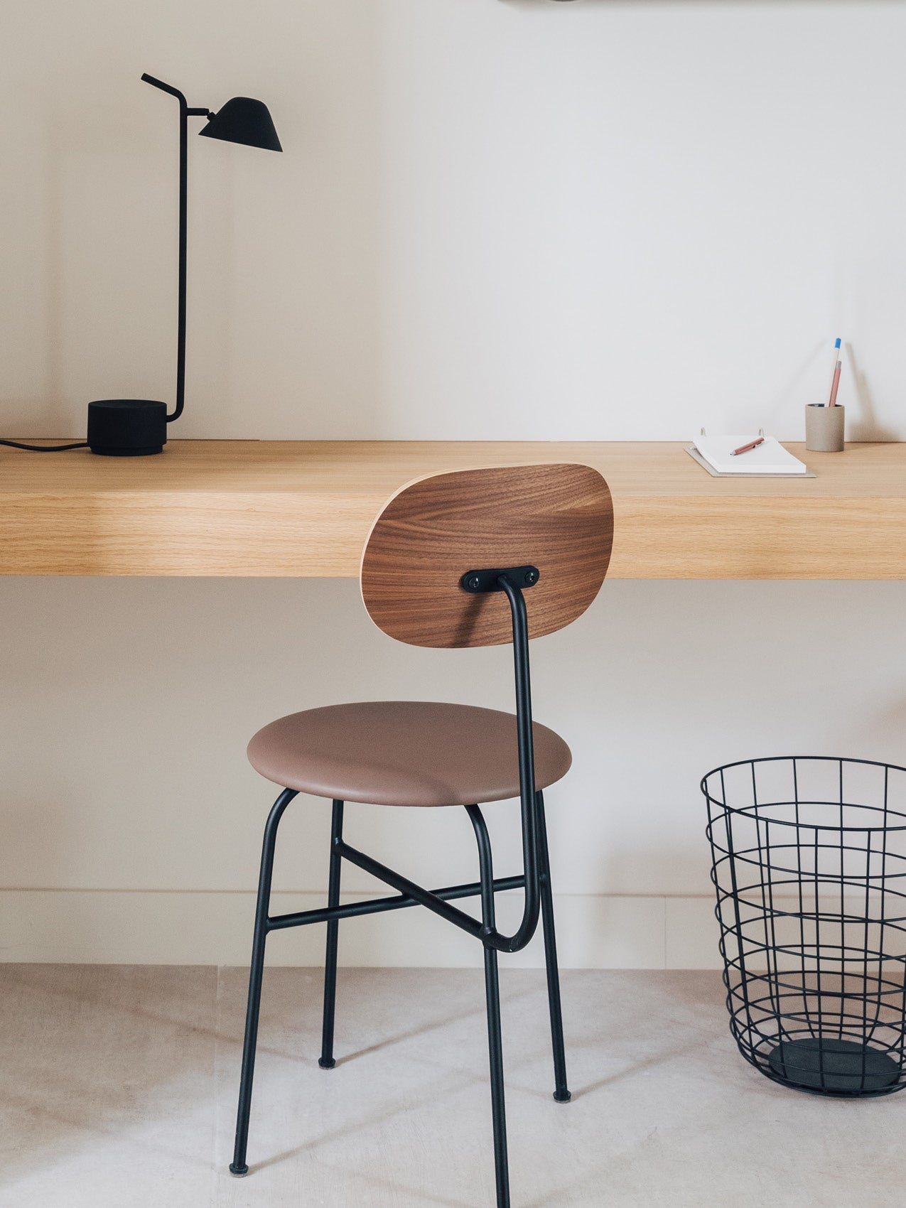 Minimal wooden desk with black metal-legged stool, brown seat and curved walnut backrest, black desk lamp and wire waste