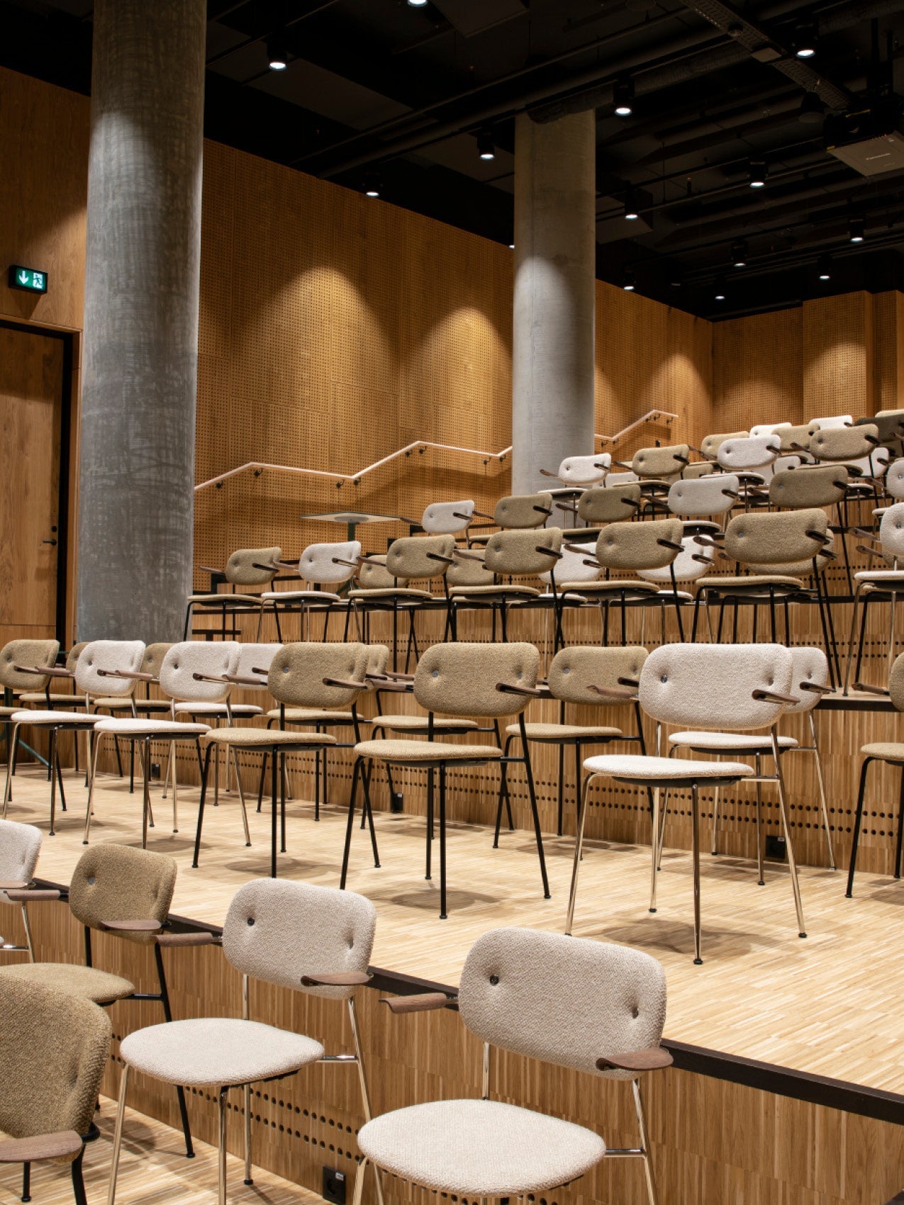 Tiered auditorium with beige wool-upholstered chairs, rounded backs and black metal legs on light wood steps.