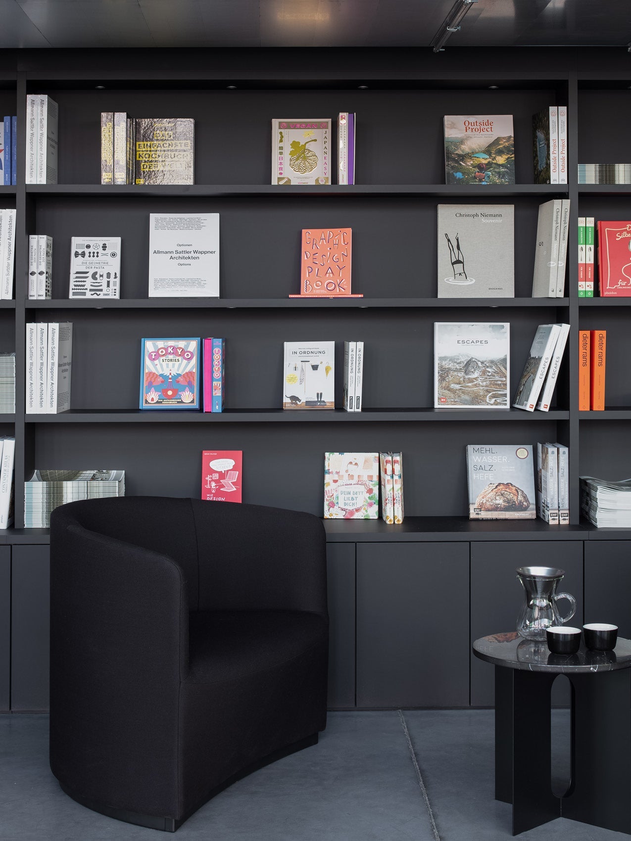 Black upholstered curved armchair beside black shelving filled with colorful books and a small black side table with