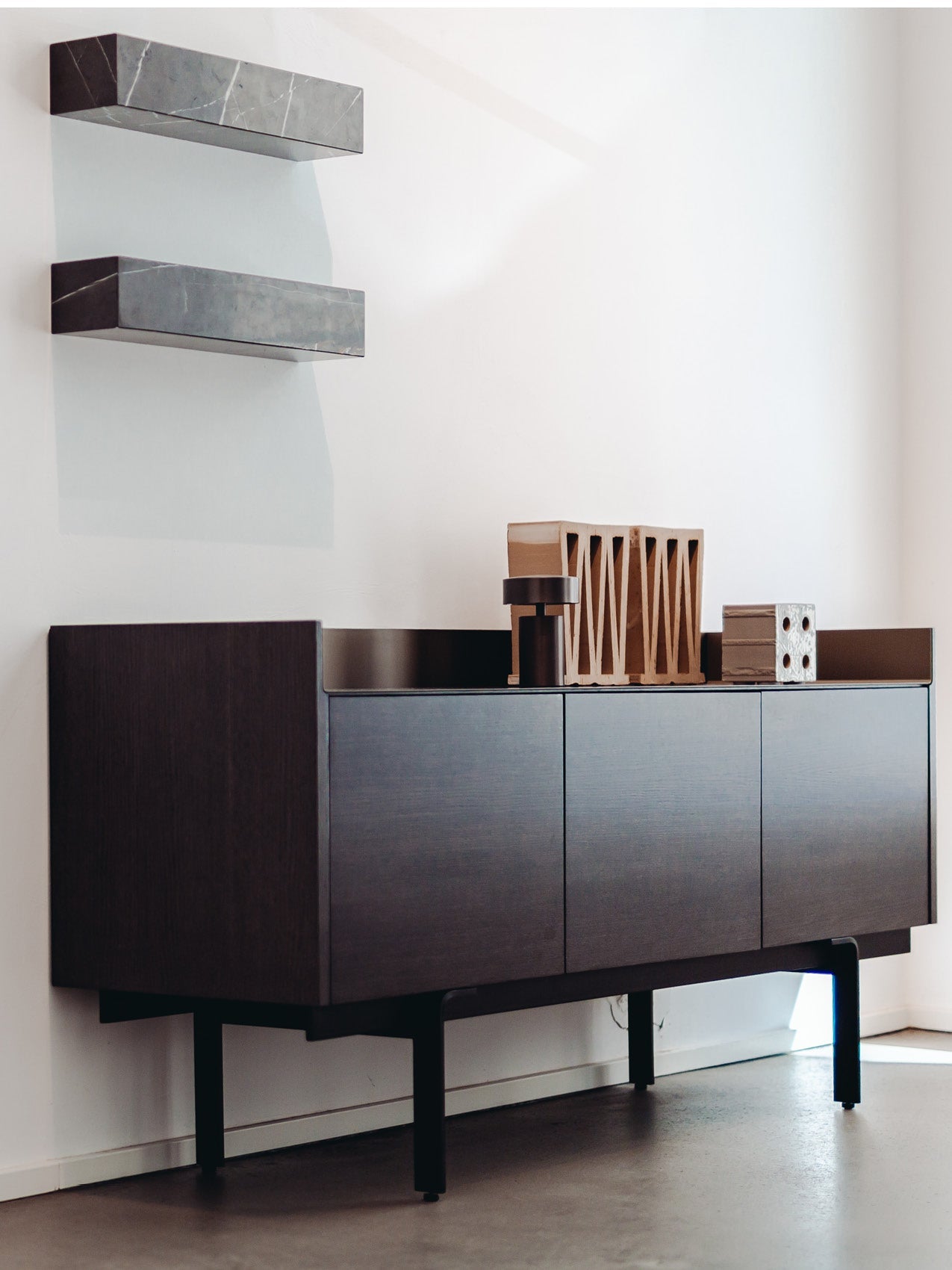 Dark wood wall-mounted sideboard with black metal legs, two floating grey stone shelves above and sculptural wooden decor on