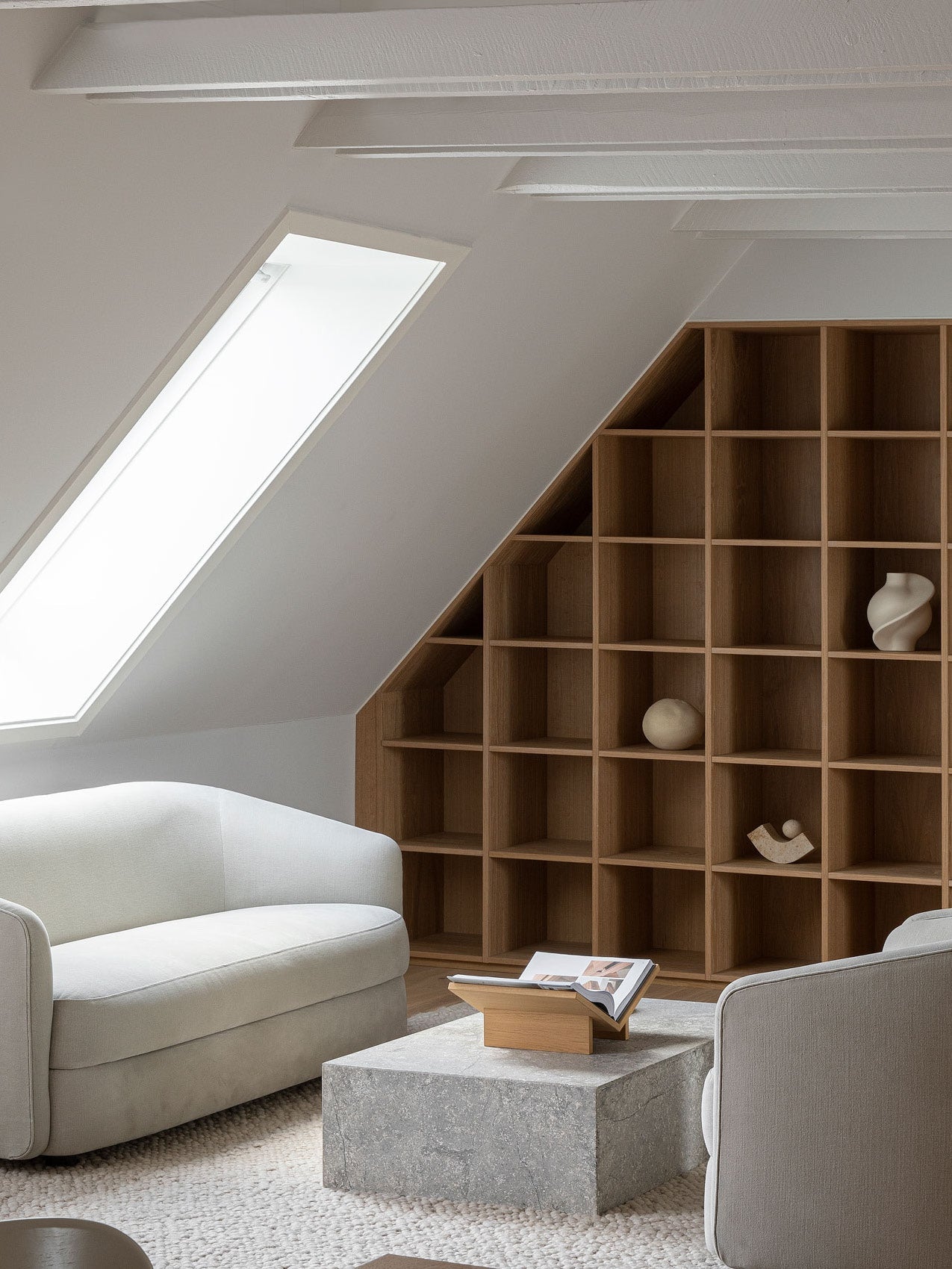 Minimal attic living room with sloped skylight, pale cream upholstered armchairs, stone coffee block and oak grid shelving.