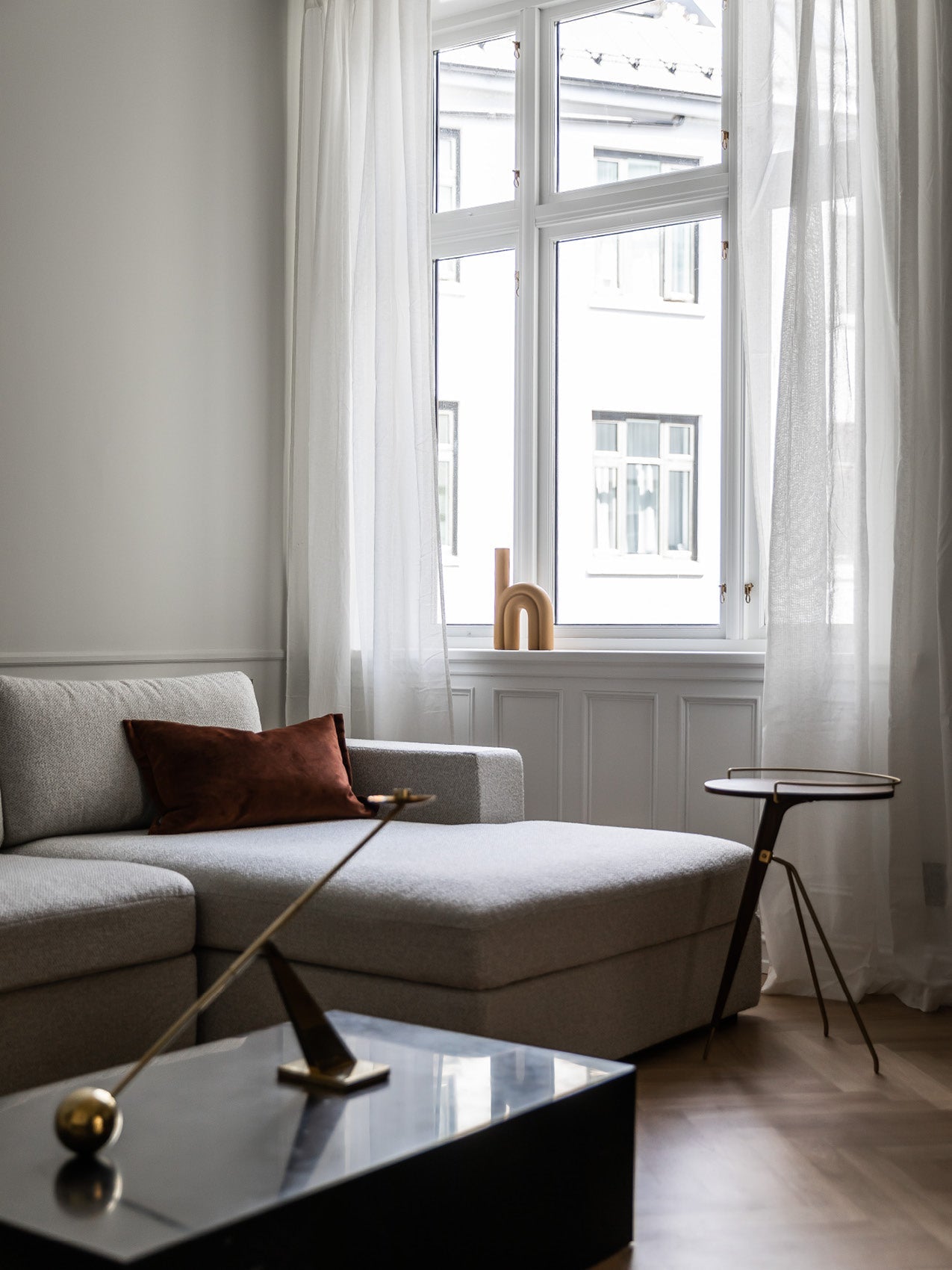 Minimal living room with light grey boucle chaise, rust velvet cushion, sheer white curtains and small wooden arch on