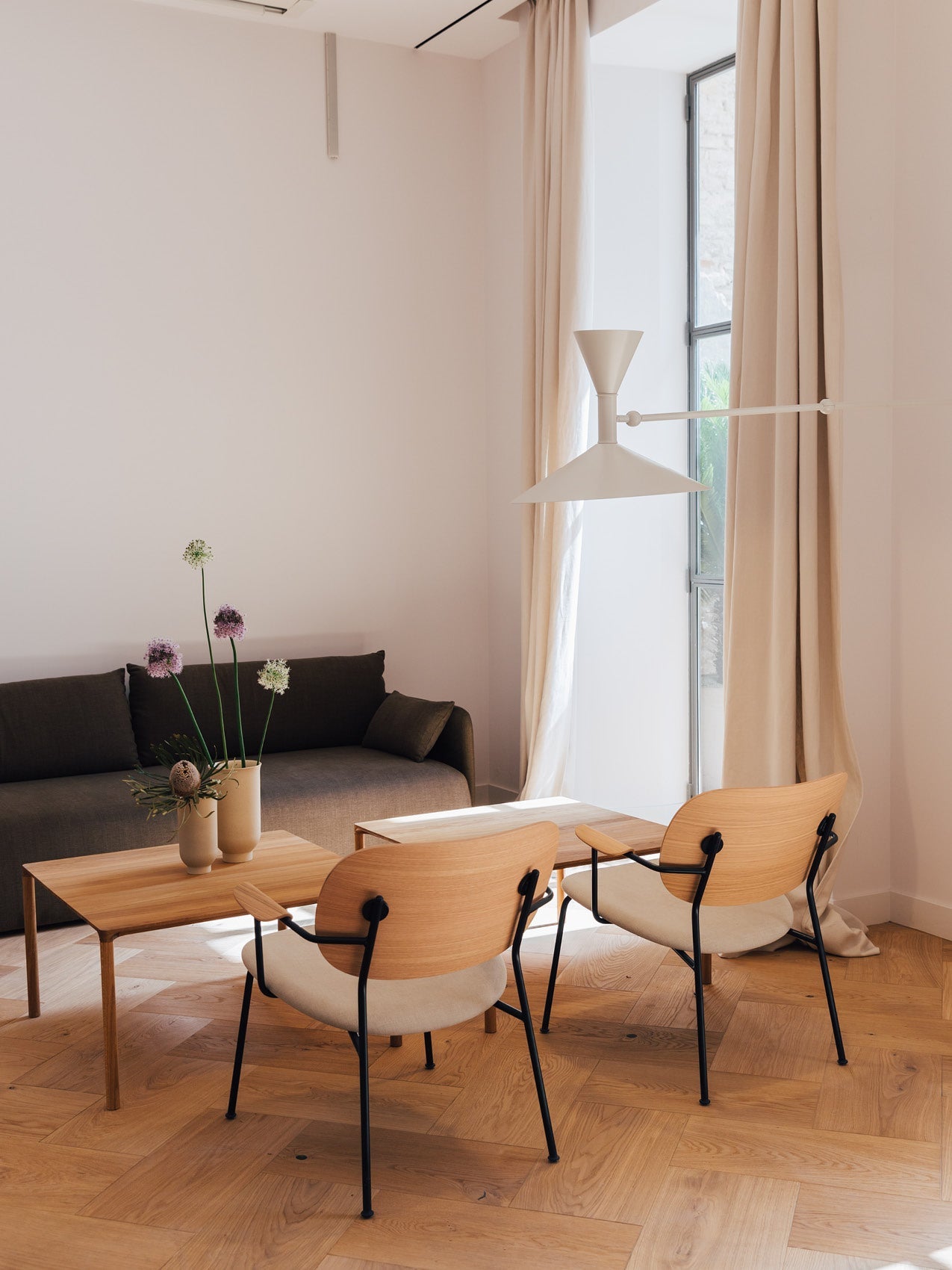 Minimal living room with light wood chairs and table, beige pendant lamp, dark grey sofa and vases with tall flowers