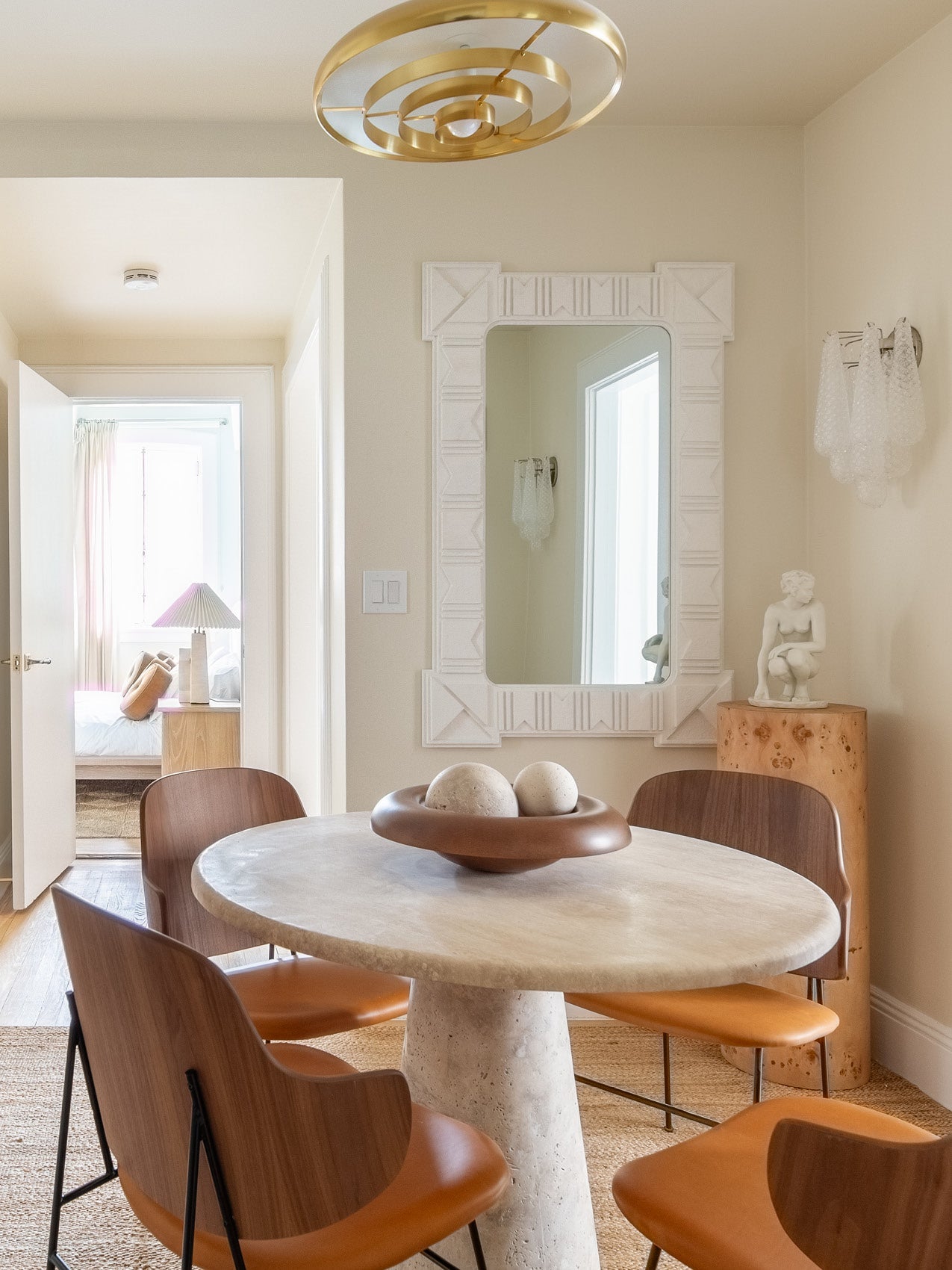 Round travertine dining table with leather-wrapped wooden chairs, terracotta ceramic bowl holding two stone spheres, gold