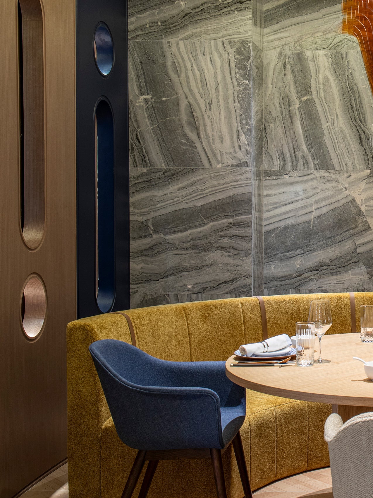 Curved mustard velvet banquette with blue upholstered chair, round wooden table set with glassware against grey veined stone