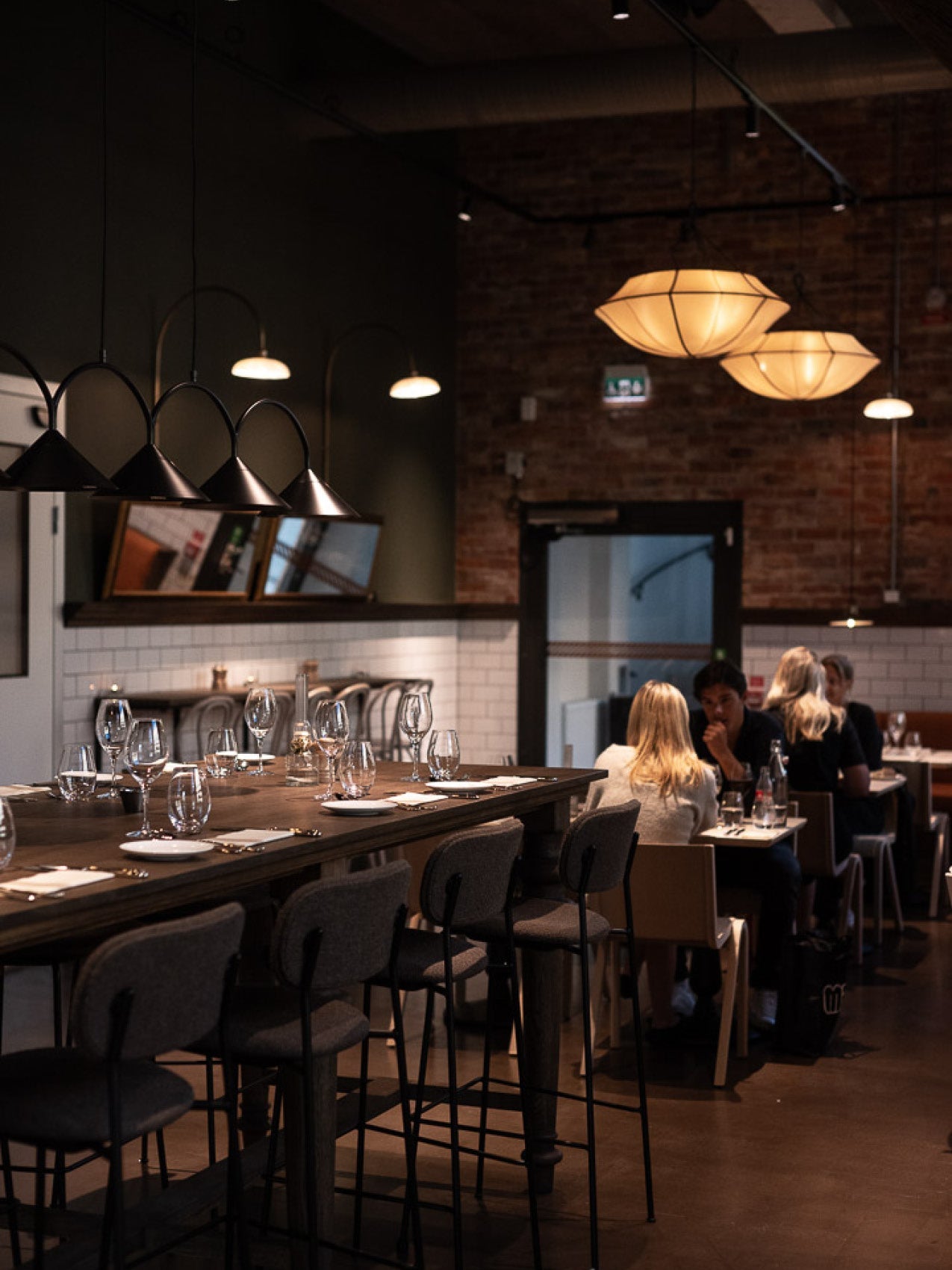 Long wooden communal table with grey upholstered bar stools, glassware and black cone and warm fabric pendant lights overhead