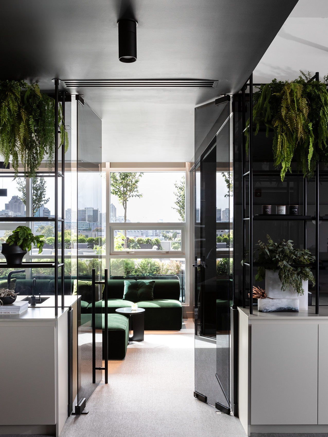 Modern office lounge with dark green velvet sectional, round black side table, glass doors and hanging ferns.