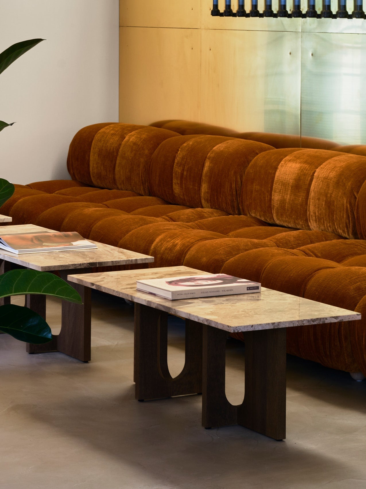 Rust-coloured tufted velvet sofa with rounded cushions beside marble-top wooden coffee tables and green plant leaves
