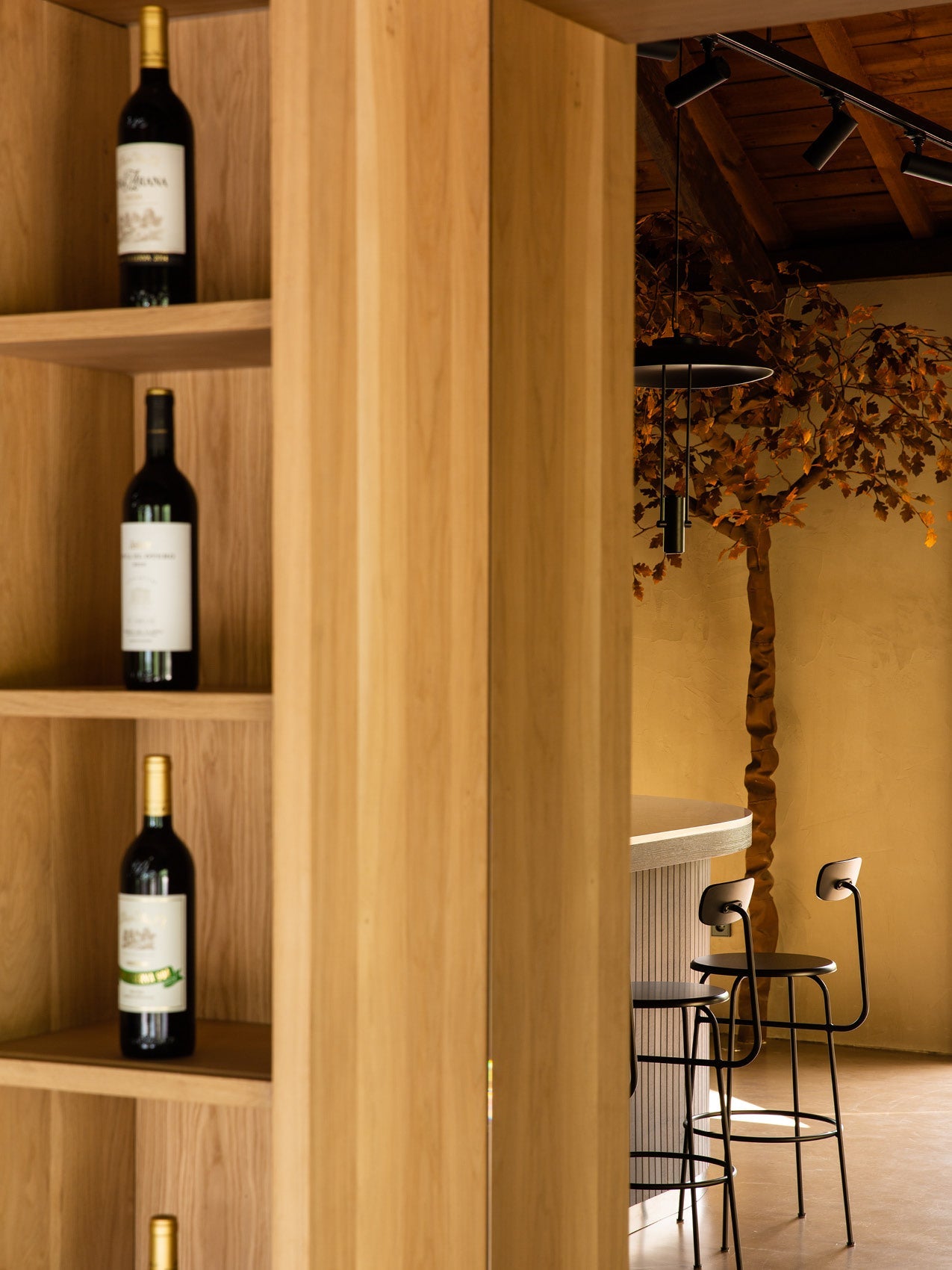 Light oak shelving with wine bottles in foreground, and black metal bar stools and pendant lamp beside a textured ochre wall.