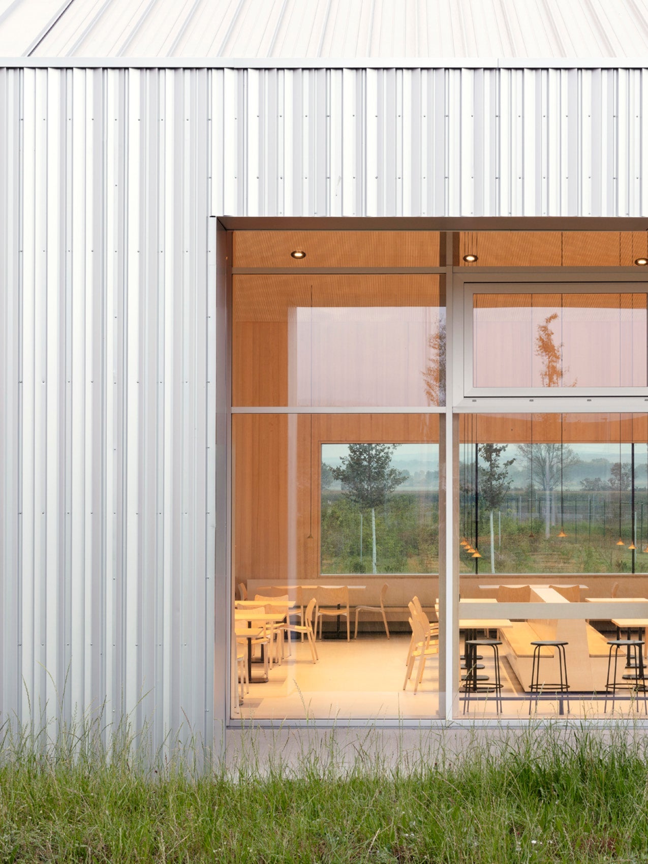 Minimalist building facade with white corrugated metal, large glass window showing warm wood interior and chairs.