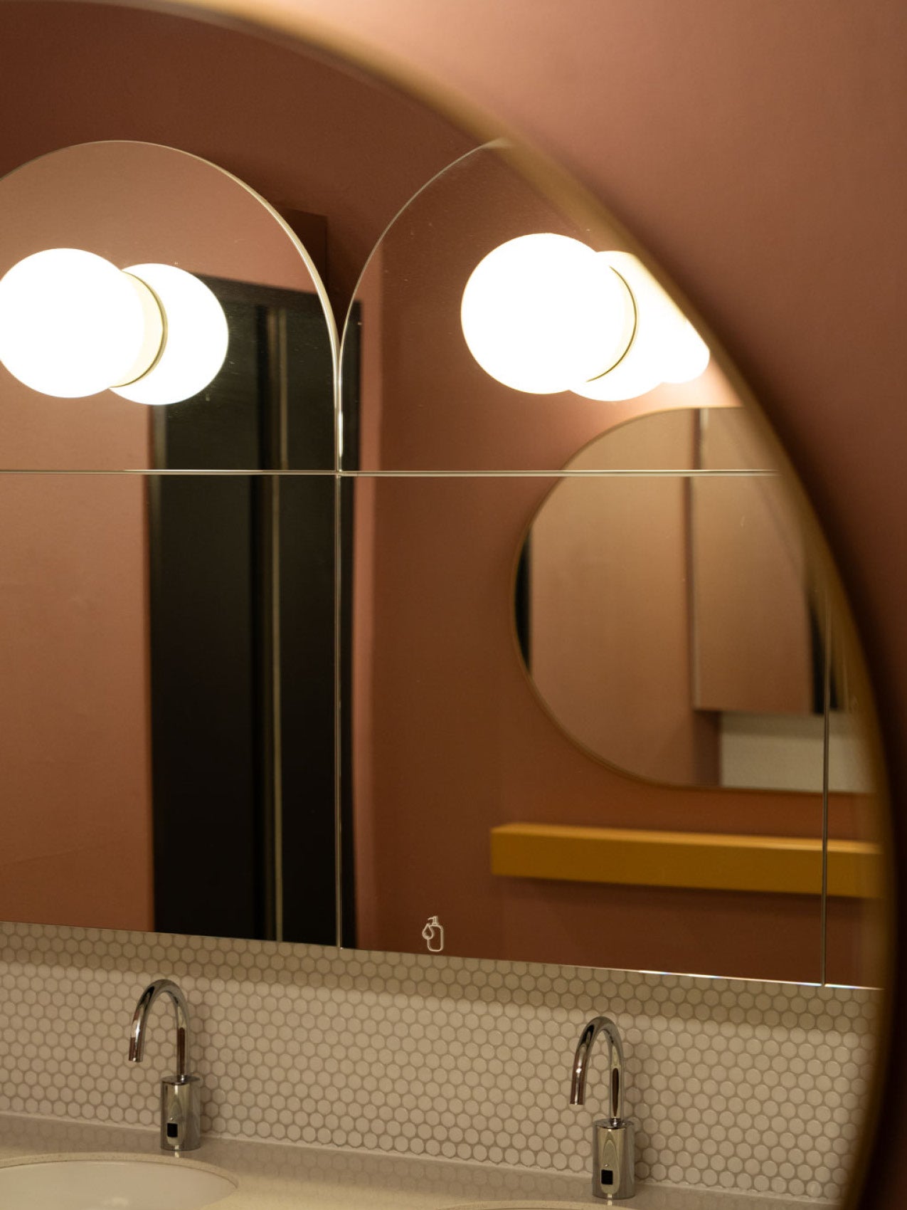 Close-up of a bathroom mirror with round frosted globe lights, terracotta wall, hexagon tile backsplash and chrome faucets
