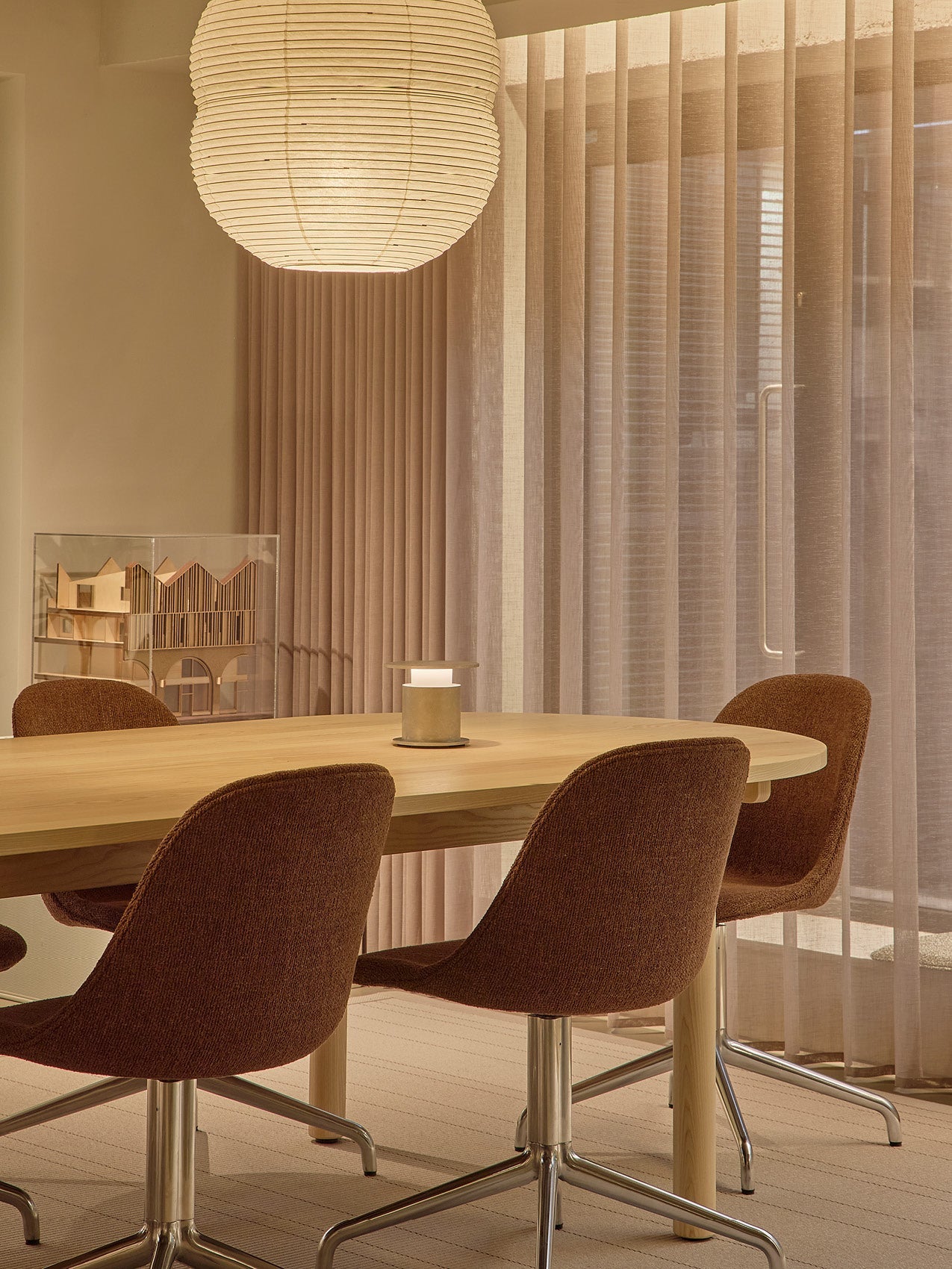 Round light wooden dining table with brown upholstered swivel chairs, paper pendant lamp and sheer beige curtains.
