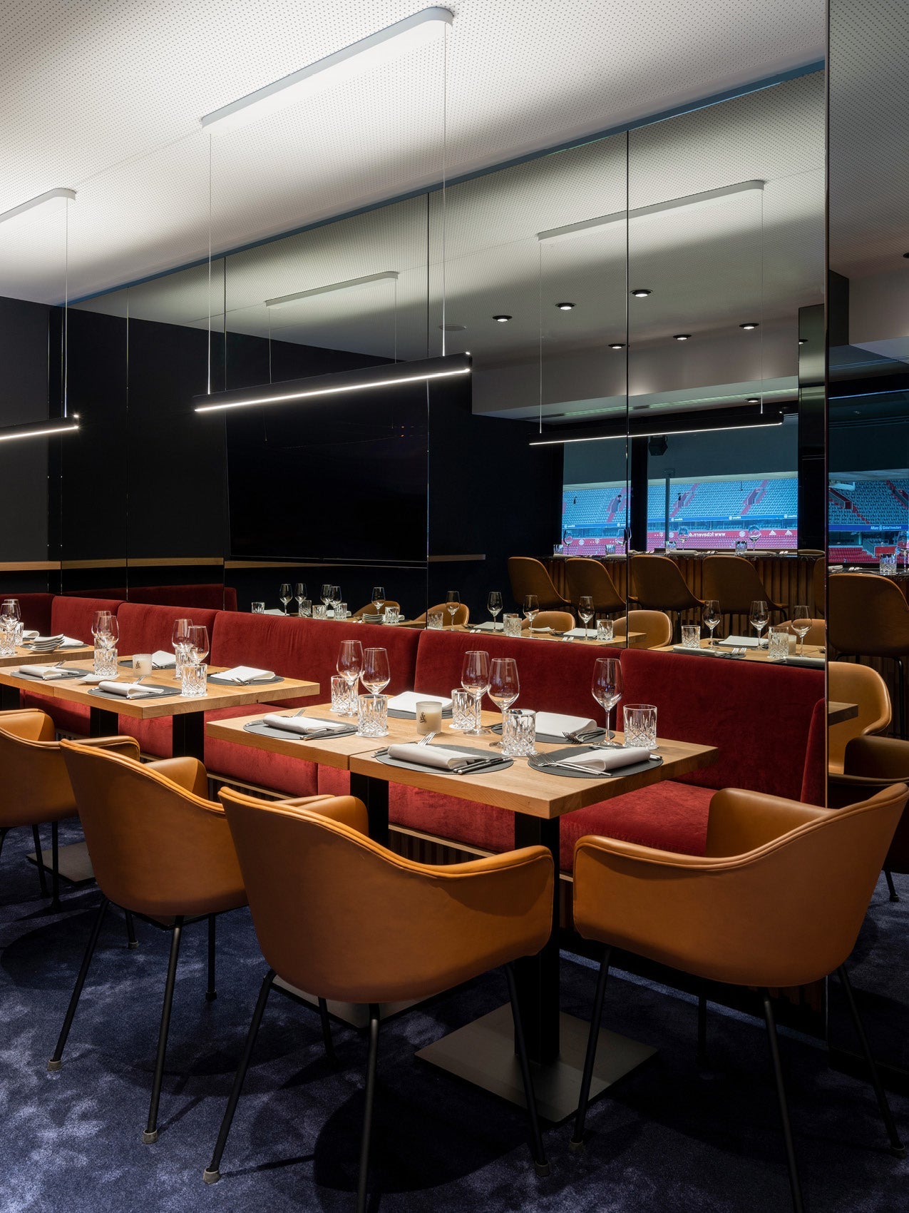 Modern dining booth with red velvet banquette, tan leather chairs, wooden tables set with glassware, and linear pendant