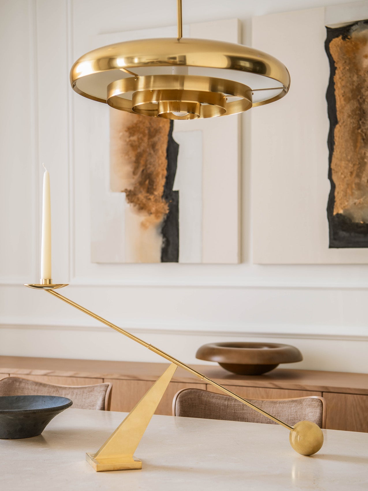 Brushed brass circular pendant light and matching angled brass candlestick with white taper on a neutral dining table