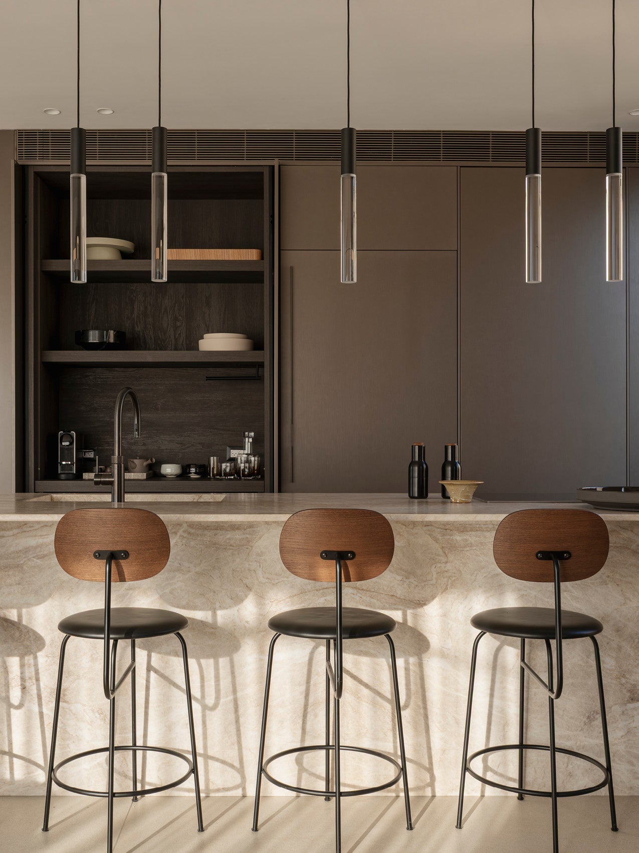 Minimal kitchen island with three wooden-back bar stools, marble countertop and pendant glass tube lights above.