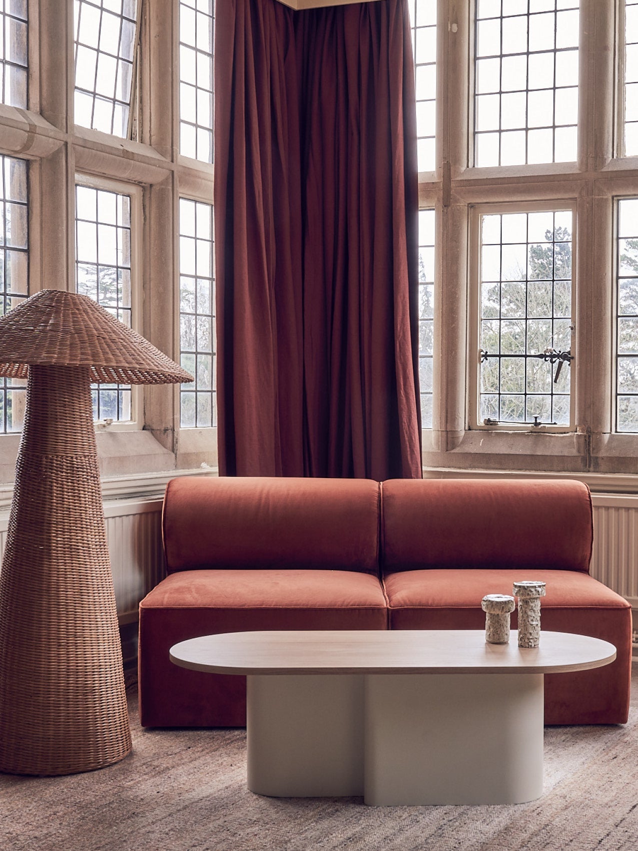 Terracotta velvet two-seater sofa with rounded cushions, cream oval coffee table, woven rattan floor lamp and tall burgundy