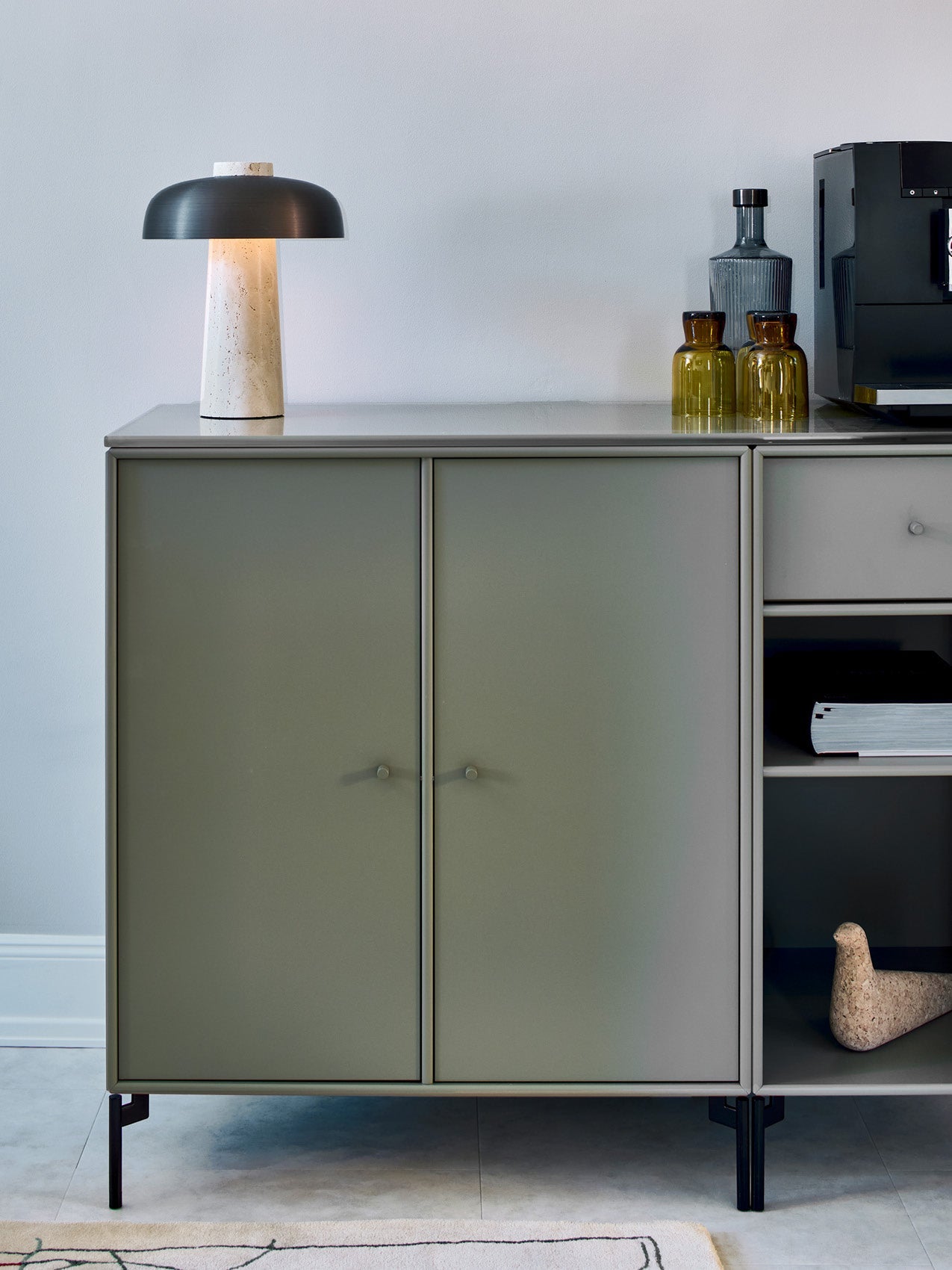 Matte olive-green metal sideboard with black legs, terrazzo-base table lamp with dark dome shade and amber glass bottles on