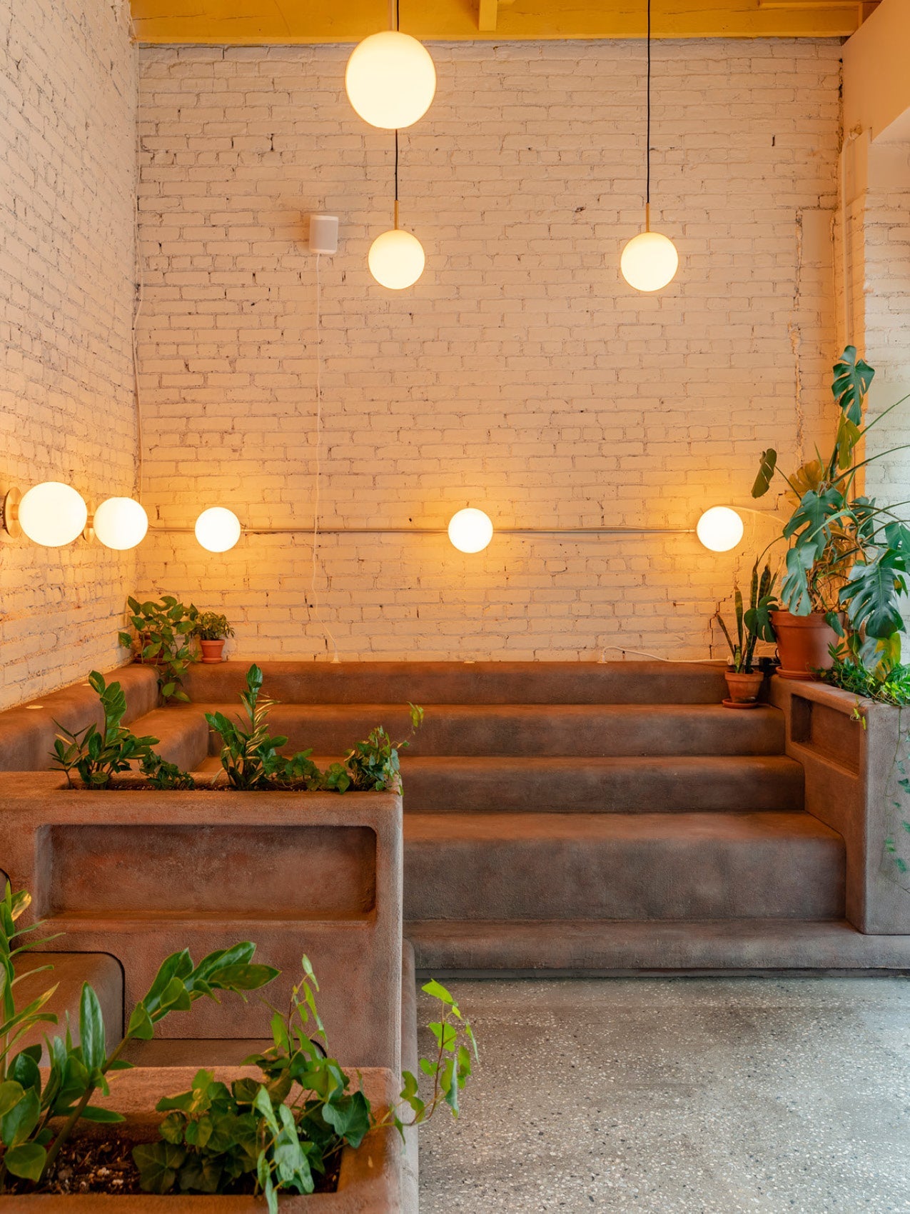 Minimalist lounge with warm globe pendant and wall lights, terracotta stepped seating and green potted plants against white