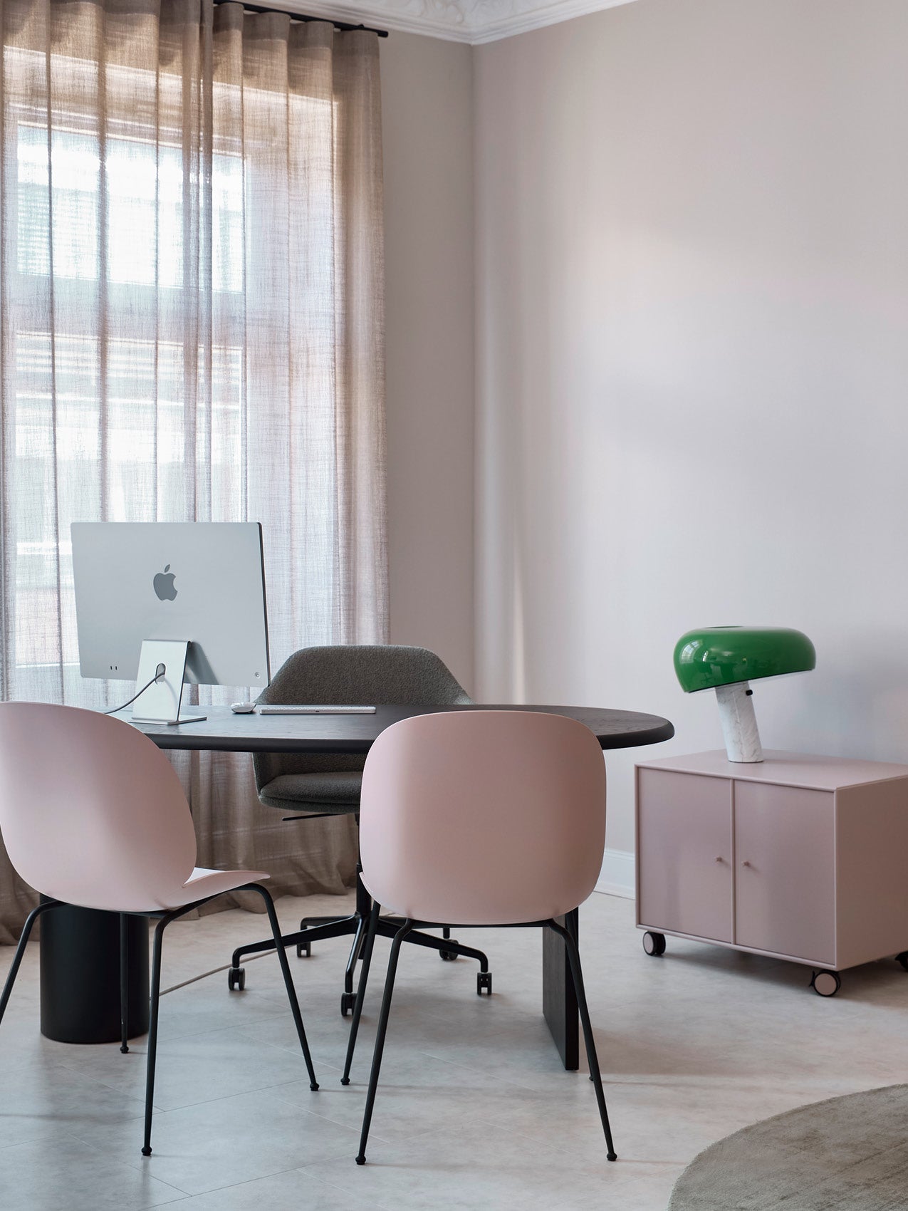 Minimal home office with round black table, pink dining chairs, grey swivel chair, iMac and green dome lamp on pink wheeled