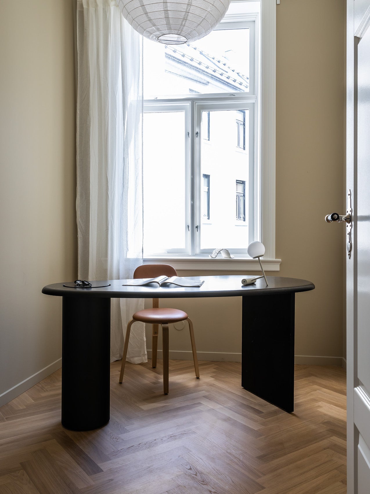 Minimal workspace with black oval table, tan leather stool, white ball table lamp and sheer curtain by a bright window