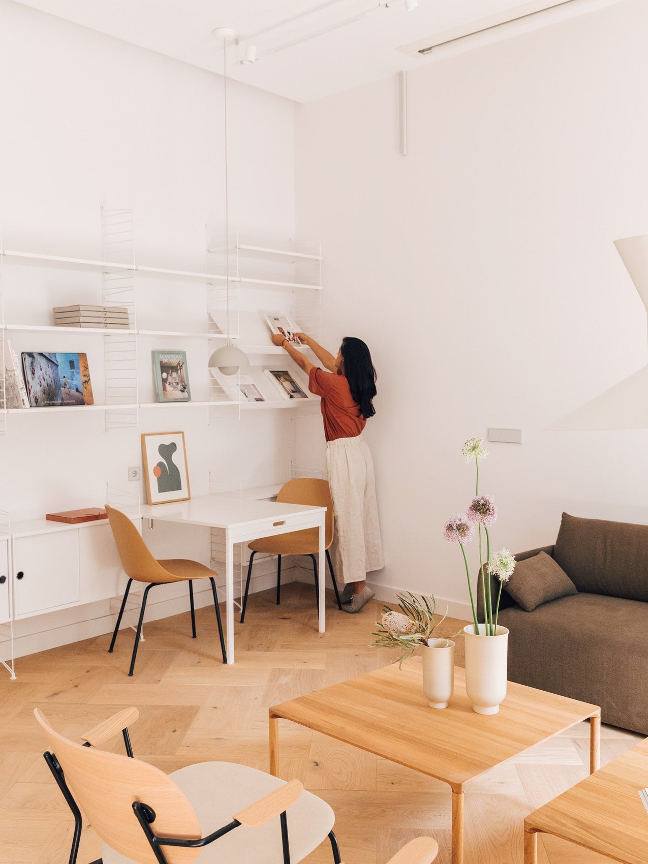 Minimal living room and workspace with white shelving, light wood furniture, tan chairs, vases with flowers, person