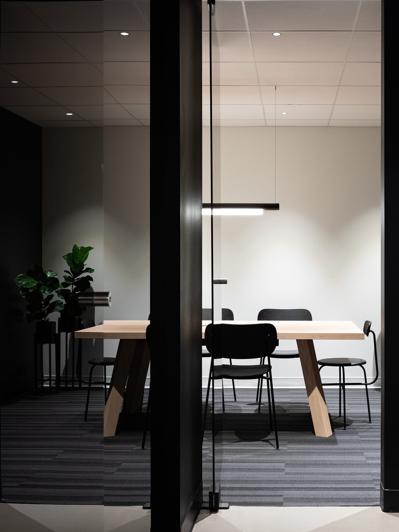 Minimal meeting room with light oak table, black chairs and linear black pendant lamp, glass door and potted plant in corner