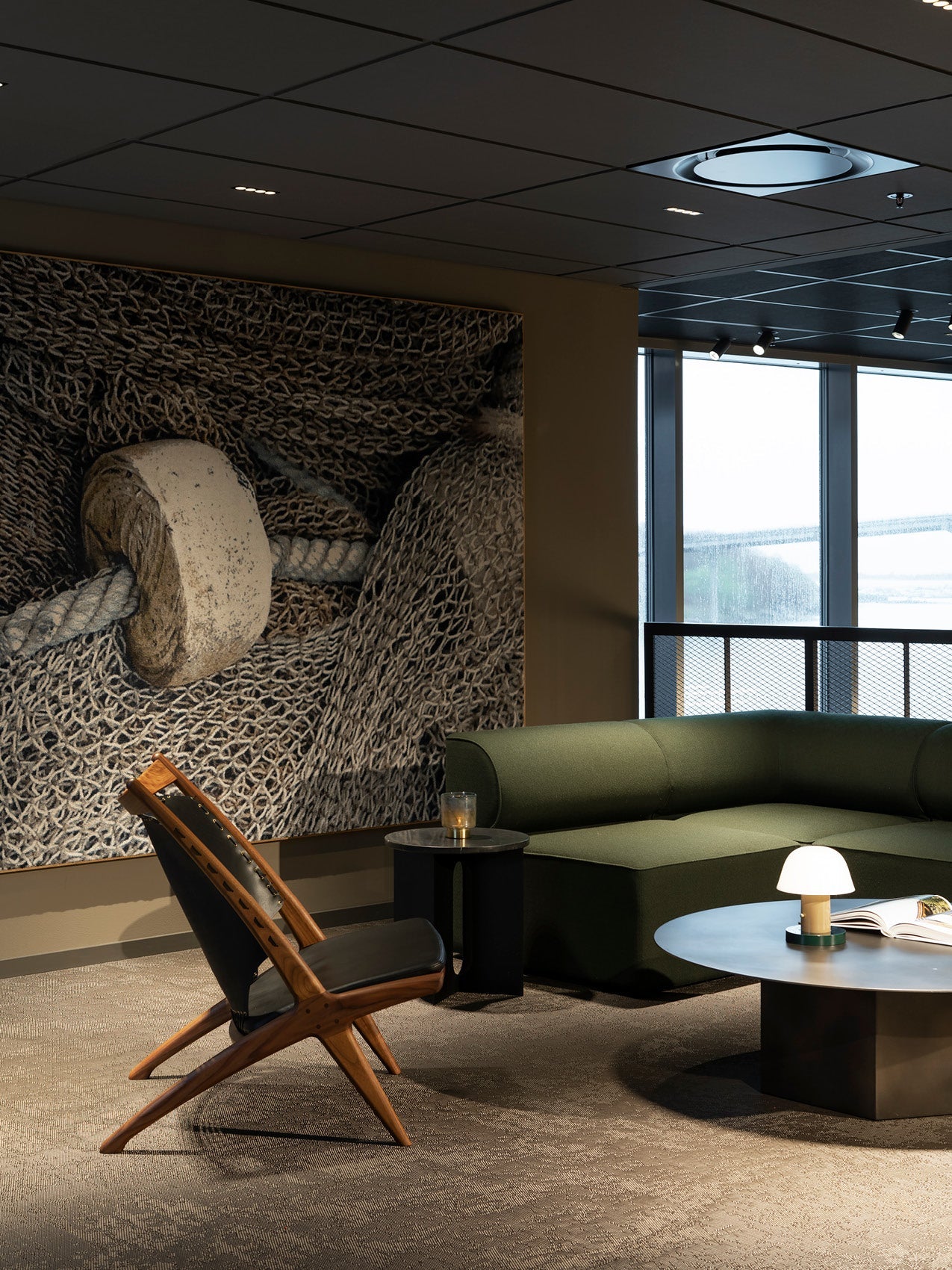 Modern lounge with dark green modular sofa, black round coffee table, wooden lounge chair and textured fishing-net wall art
