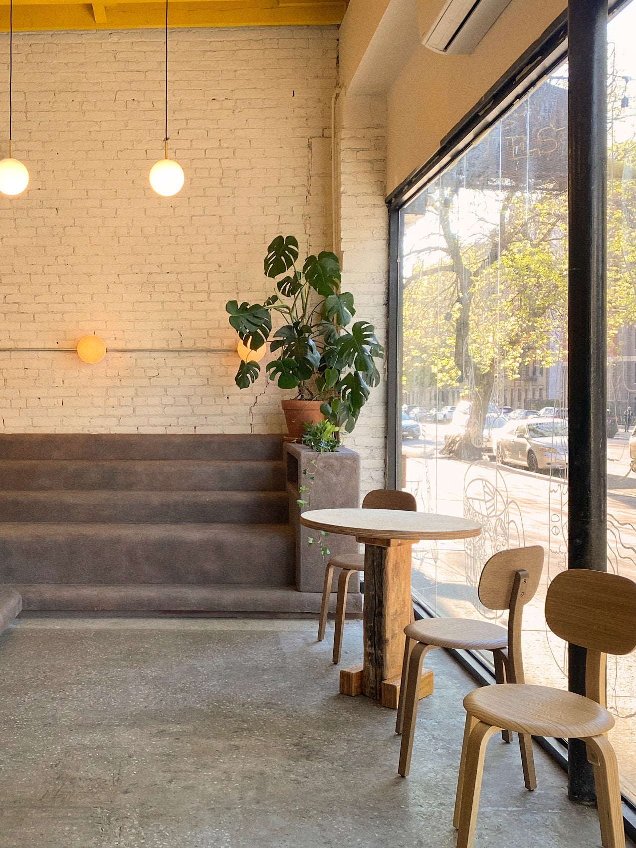 Minimal café interior with round wooden table and bentwood chairs by large window, beige brick wall, globe pendant lights