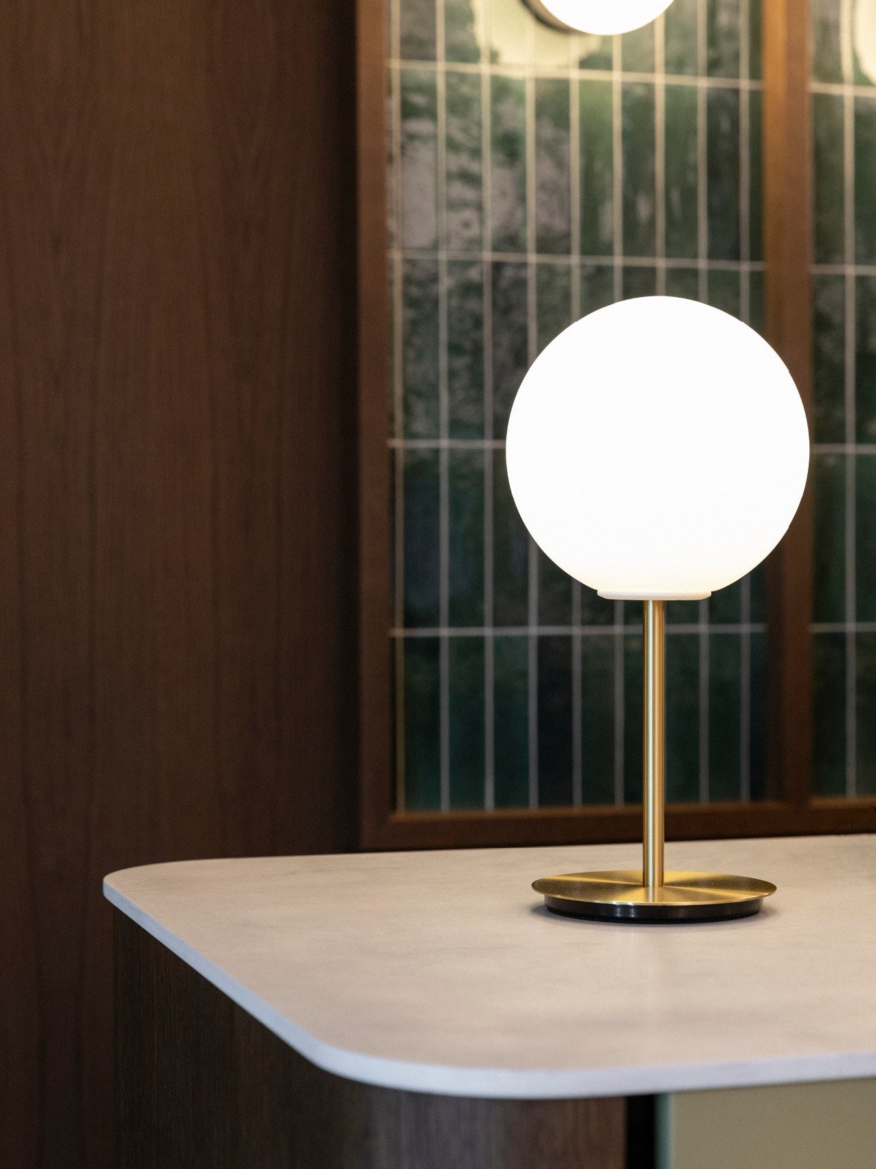 Brass table lamp with frosted white glass globe on a marble-topped counter, dark wood and tiled window background
