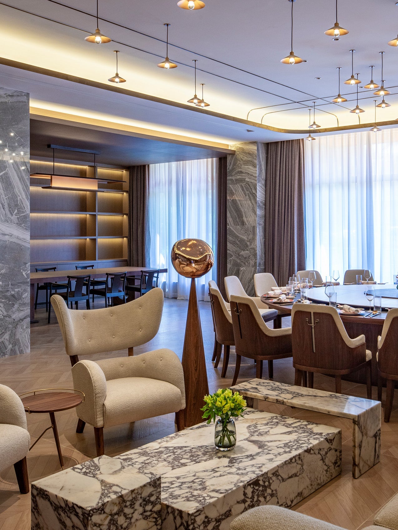 Luxurious dining and lounge area with beige upholstered chairs, marble low tables, wooden round table, draped windows and